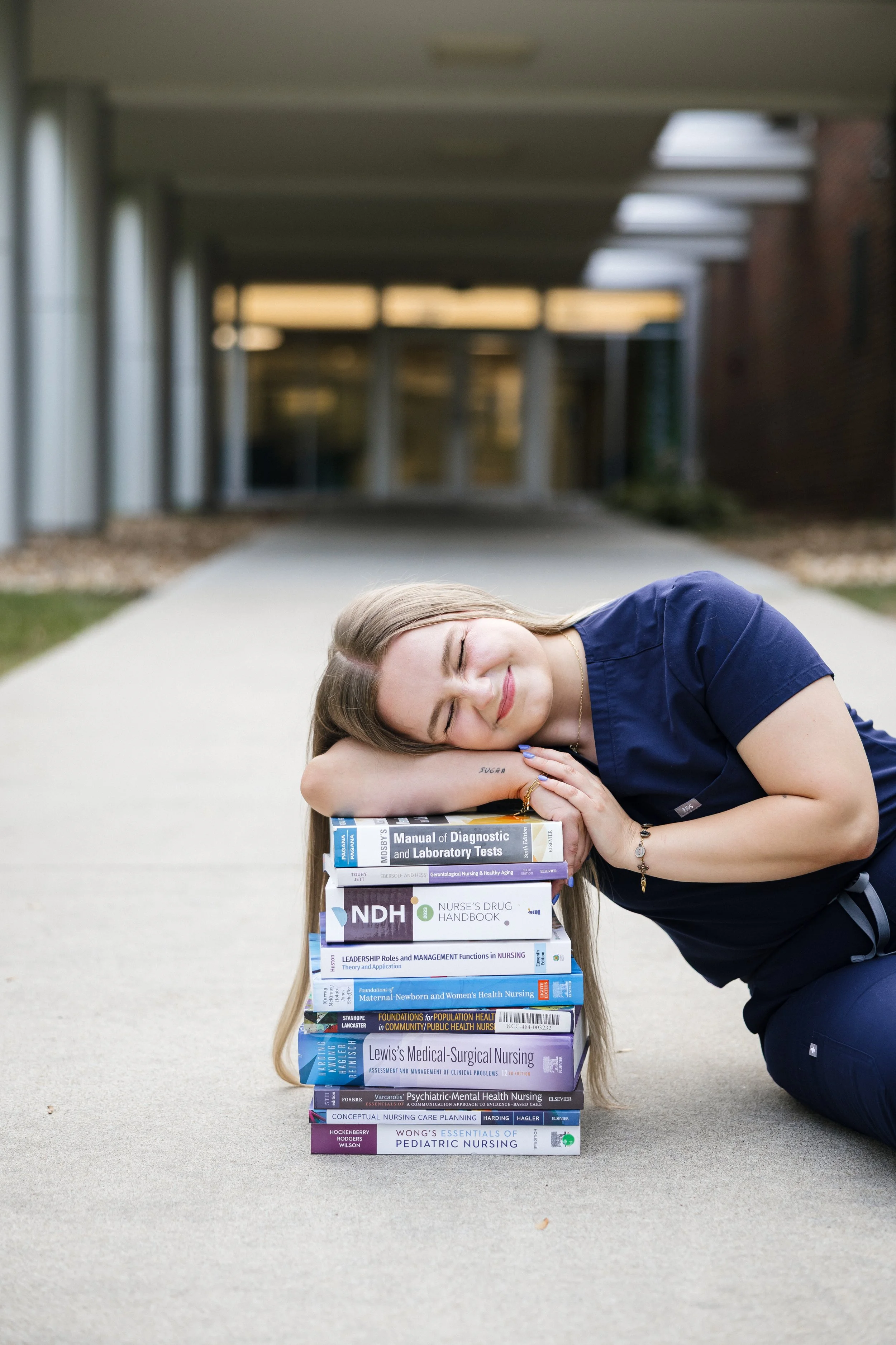 Young woman in navy scrubs resting her head on a stack of nursing textbooks on a sidewalk outside a building.