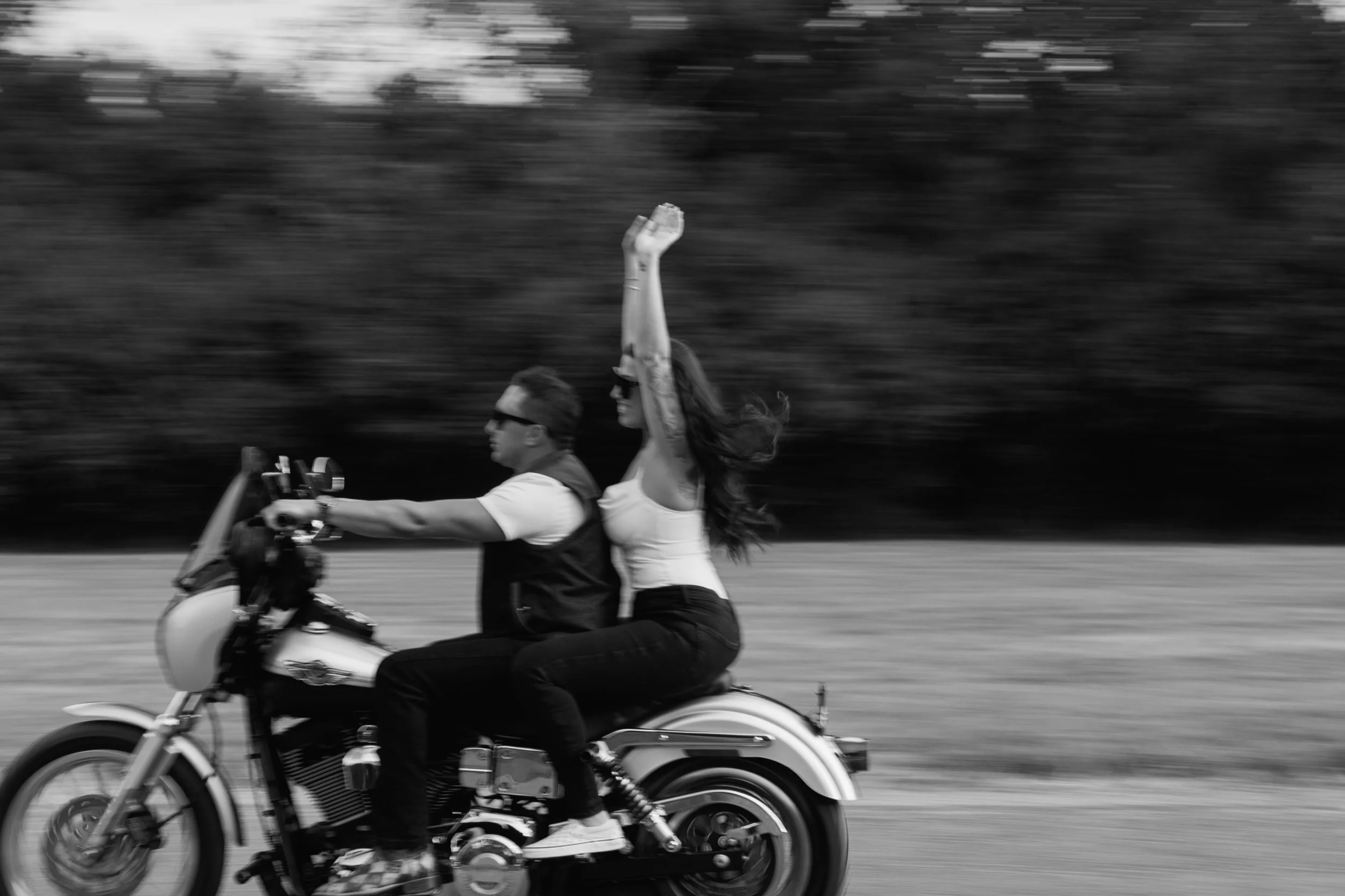 Black and white photo of a woman with long wavy hair laughing and a man in the background smiling in an open field with trees in the distance.