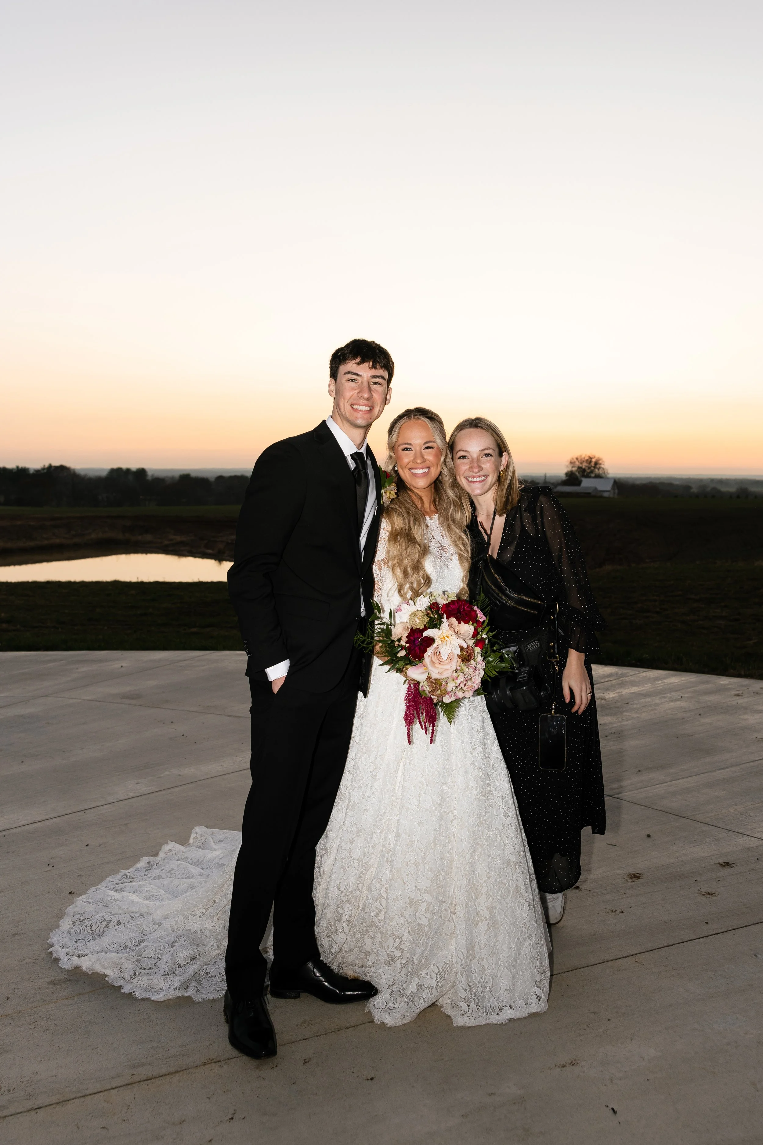 A newlywed couple stands outdoors during sunset, with a woman holding a bouquet of flowers and a photographer girl beside them.