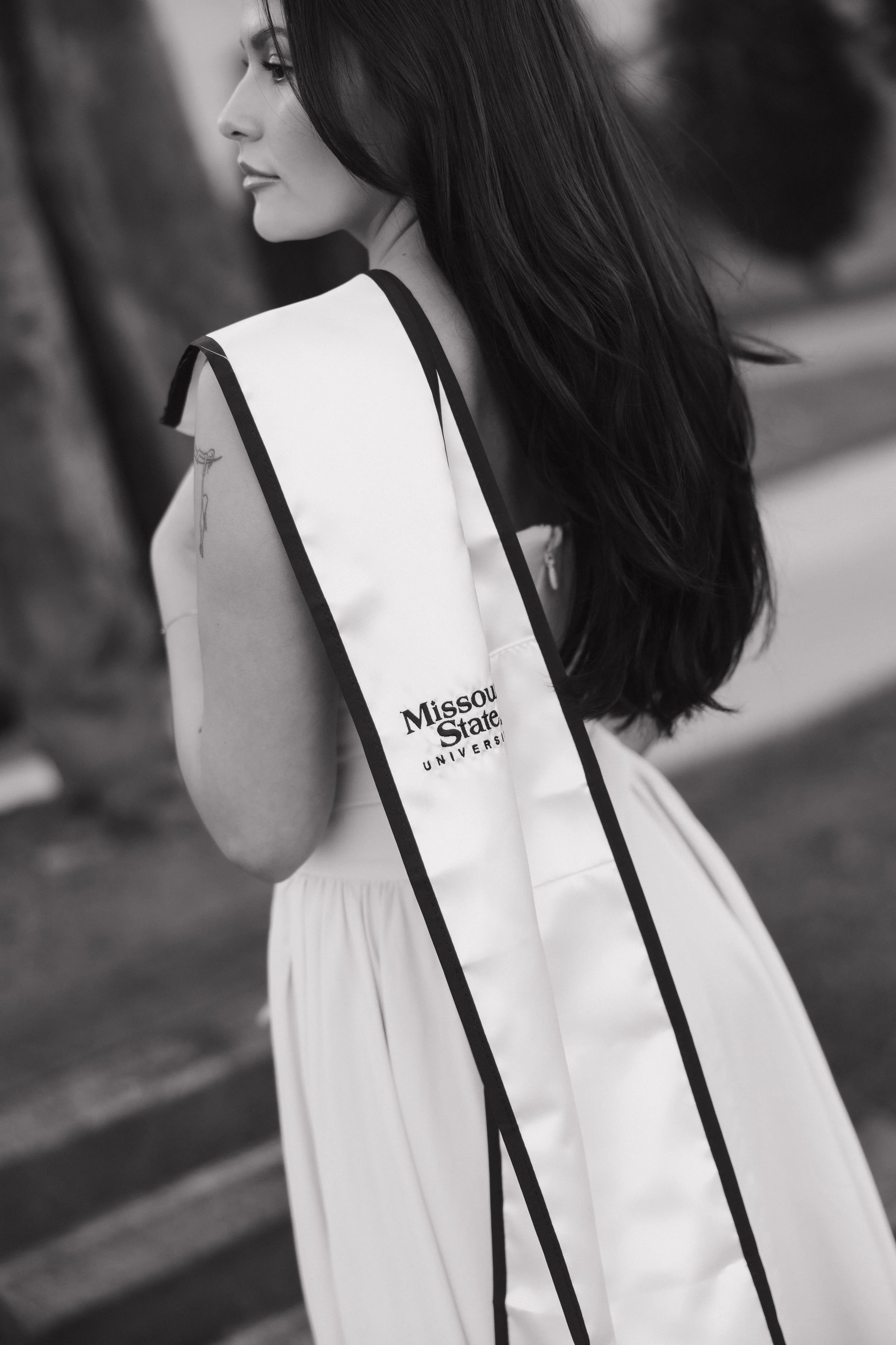 A woman with long dark hair wearing a white dress and a sash that reads 'Missouri State University' is outdoors near trees.