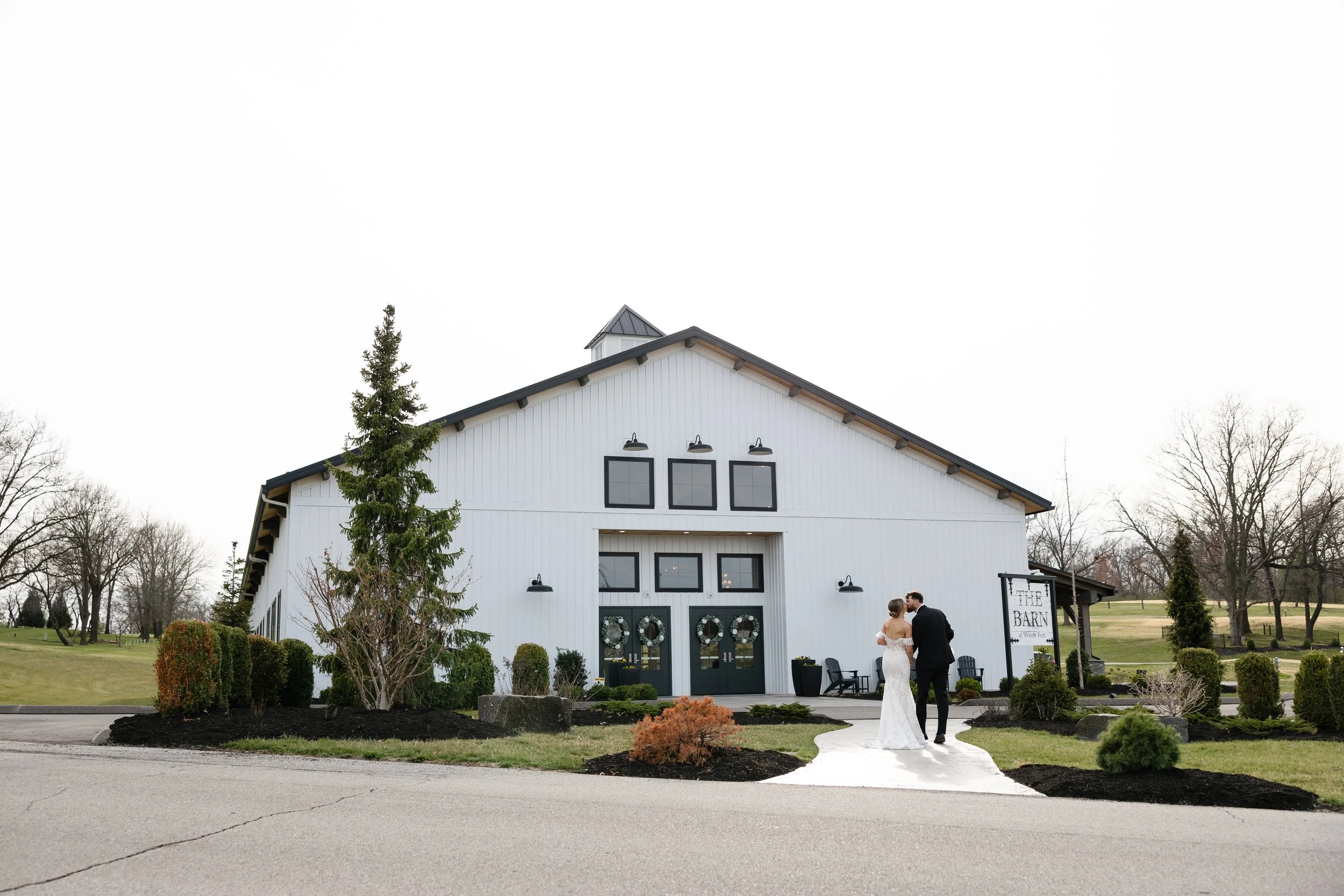 A bride and groom stand outside a white barn decorated for a wedding, with the bride in a white dress and the groom in a black suit, holding hands and looking at each other.