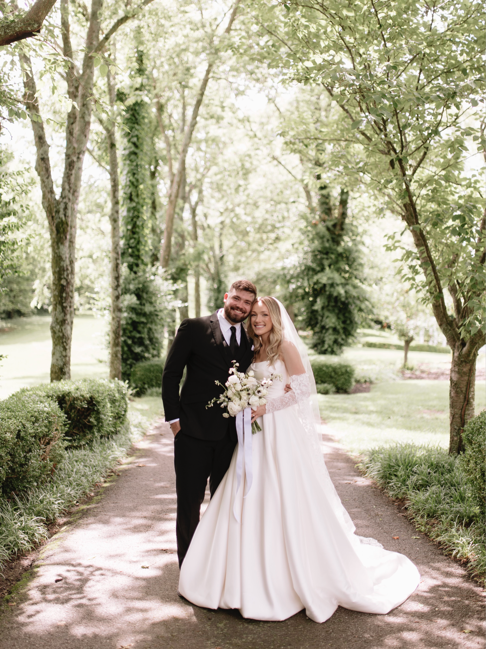 A bride and groom standing on a sunlit path surrounded by trees, smiling, with the bride holding a bouquet of white flowers.