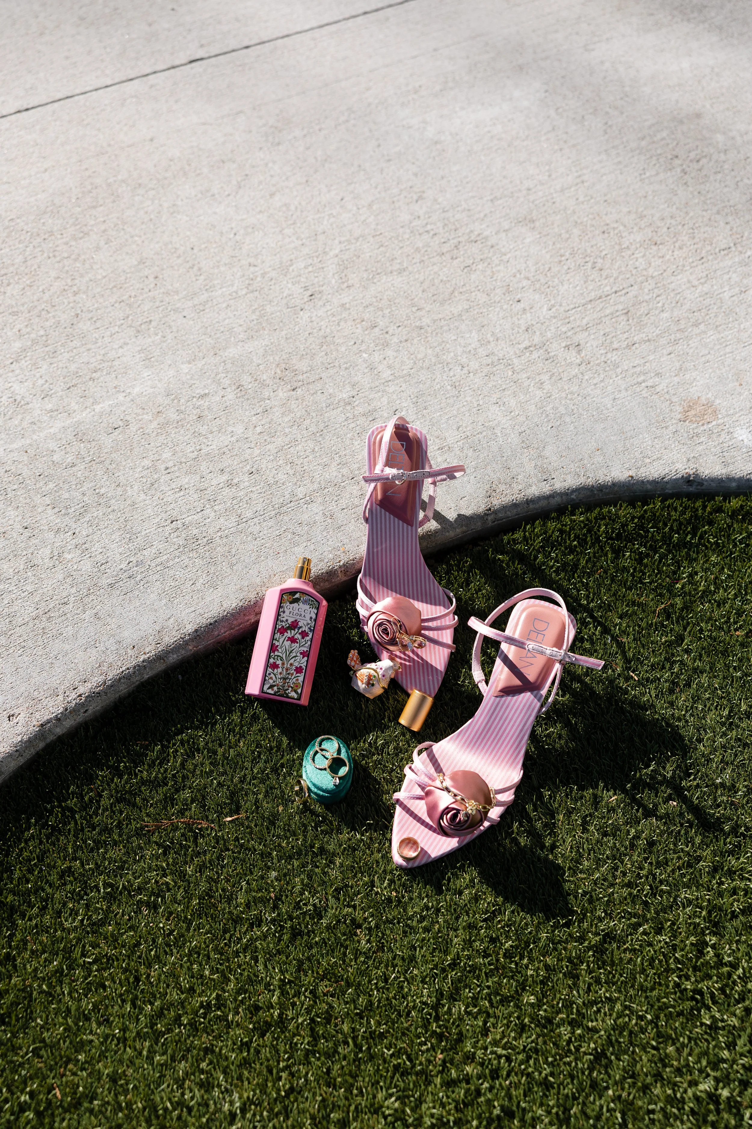Pink and white striped high-heeled shoes, a pink perfume bottle with floral design, a turquoise clutch with rings, and a small cosmetics container, all arranged on a grassy area near concrete pavement.