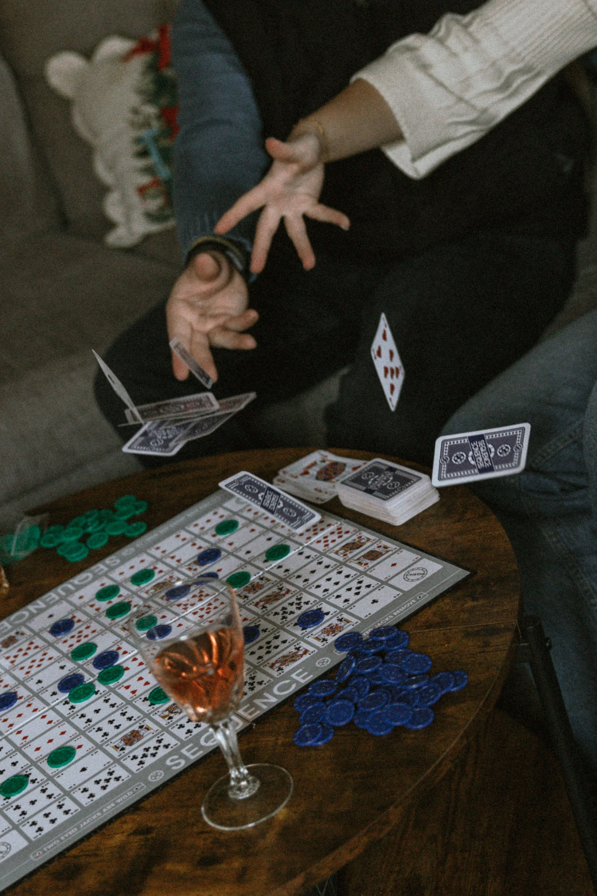 People playing a game of Rummikub on a wooden table, with poker chips, playing cards, a glass of rosé wine, and a hand of dominoes visible.