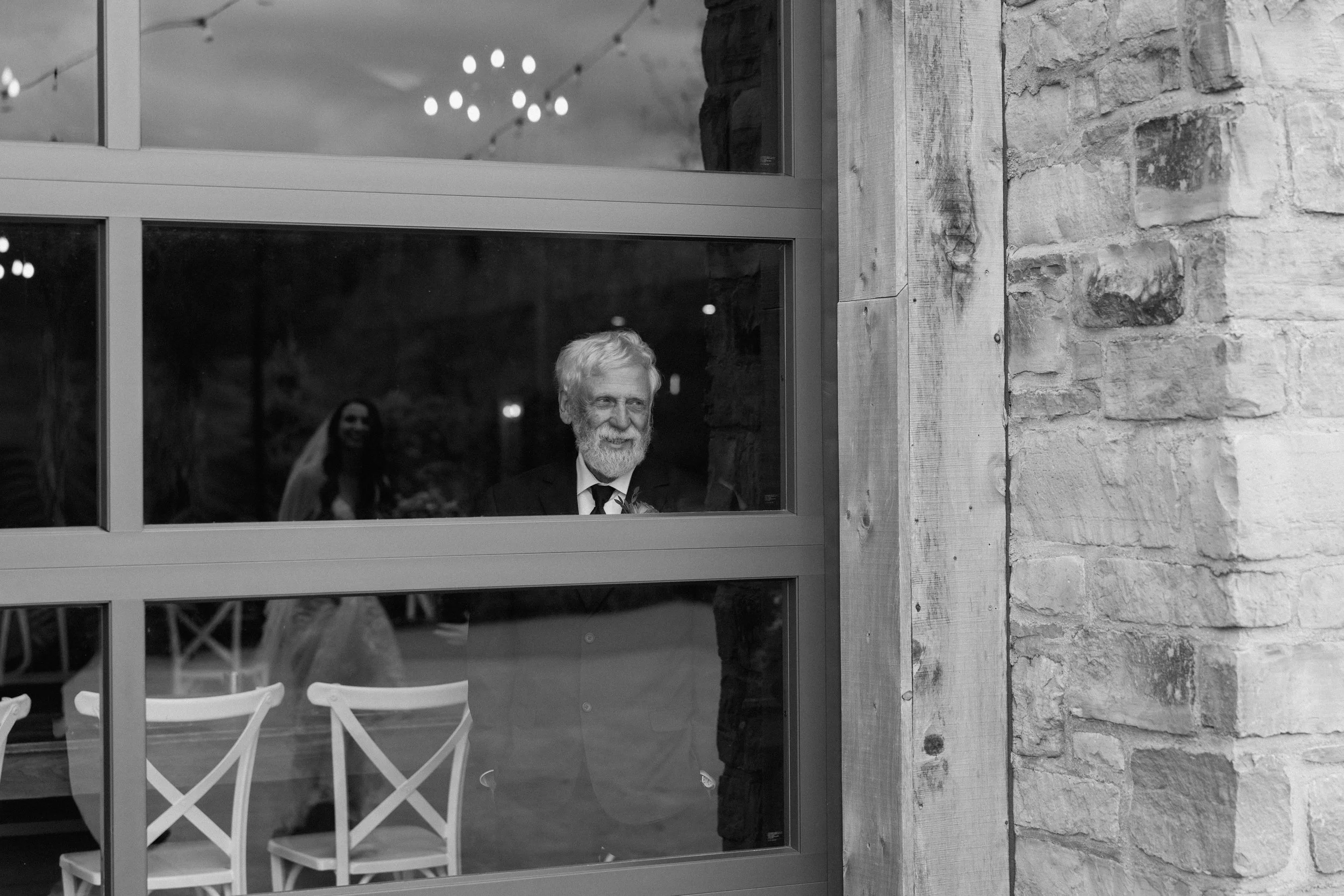 An elderly man with a beard dressed in a suit and tie looking through a window, with a smiling bride in a wedding dress and veil visible in the reflection behind him.
