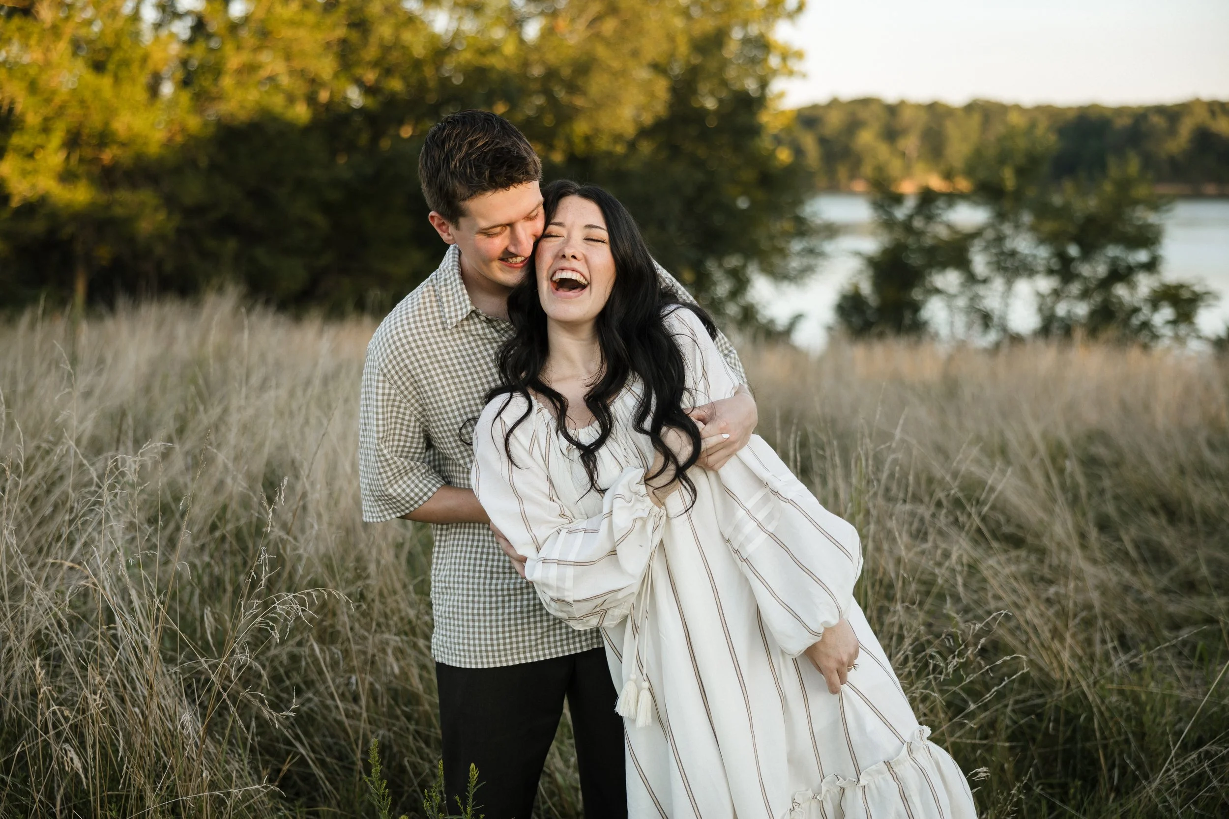 A joyful couple laughing and embracing outdoors in a grassy field with trees and a body of water in the background during sunset.