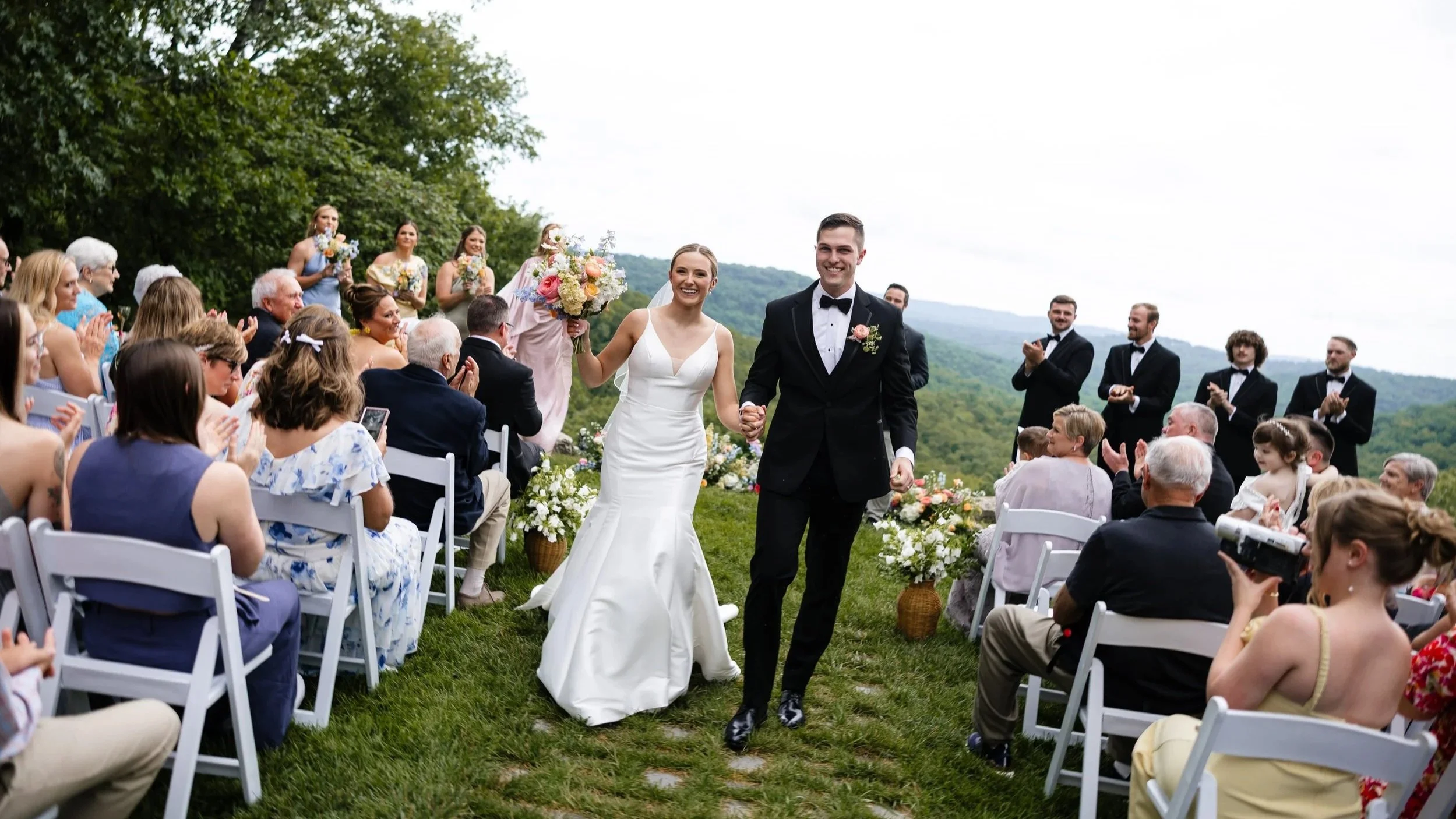 A bride and groom facing each other during their wedding ceremony outdoors among trees, with the groom reading vows from a small book.