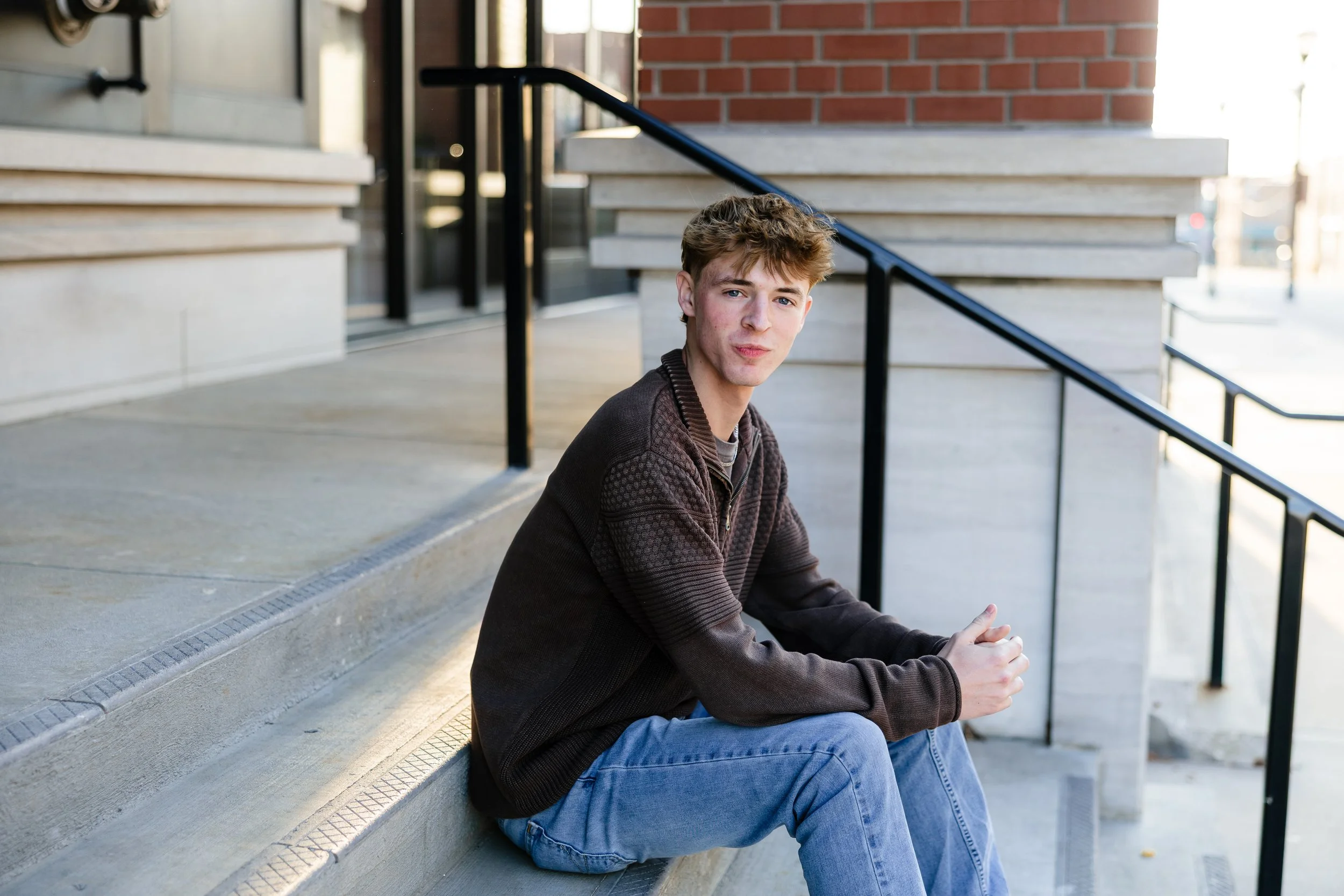 A young man with light brown hair and fair skin sitting on concrete steps outdoors near a building with brick and beige walls. He is wearing a dark brown sweater and light blue jeans, with hands clasped together.