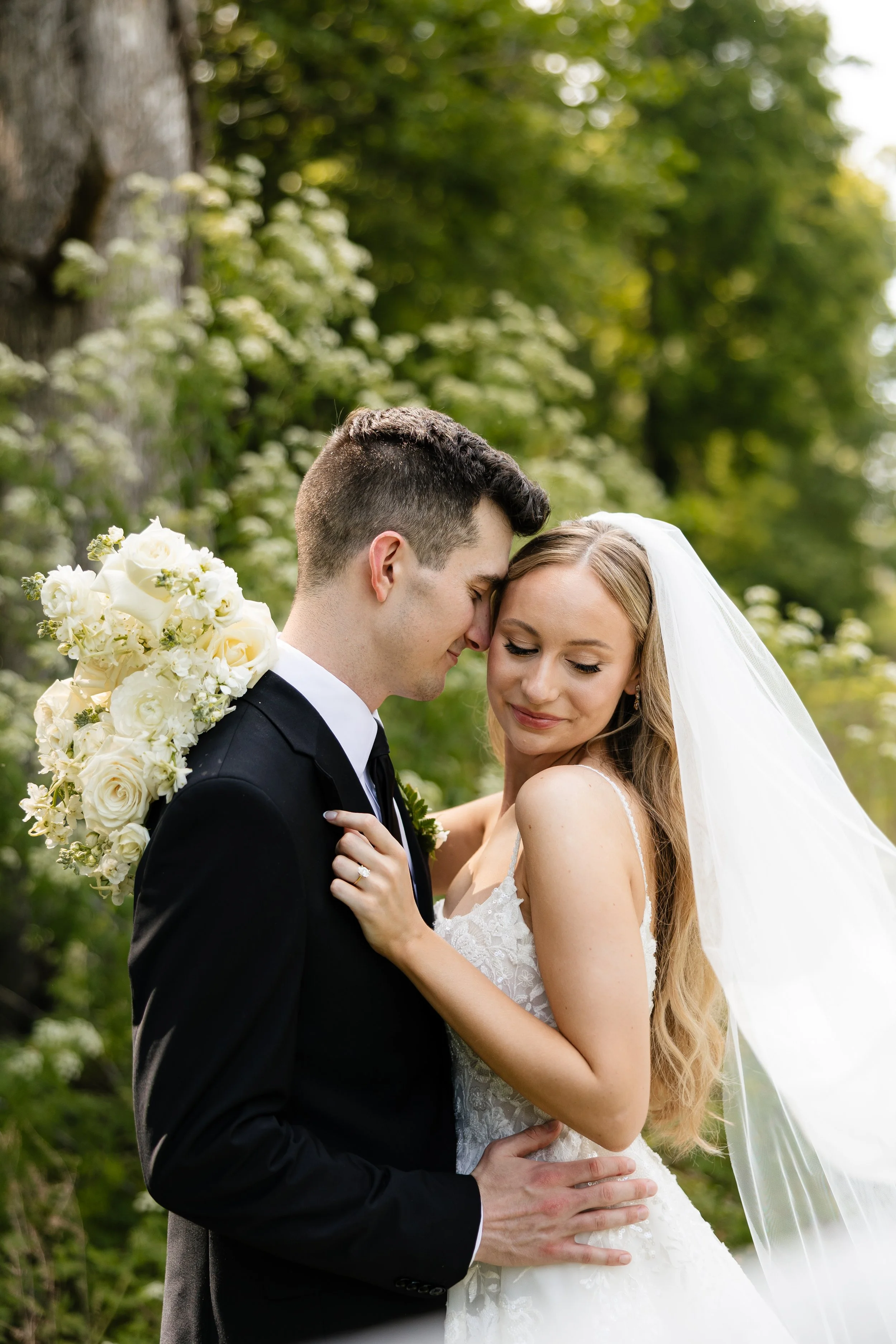 A newlywed couple embraces outdoors; the groom holds a bouquet of white and cream flowers over his shoulder, and their foreheads touch as they smile gently.