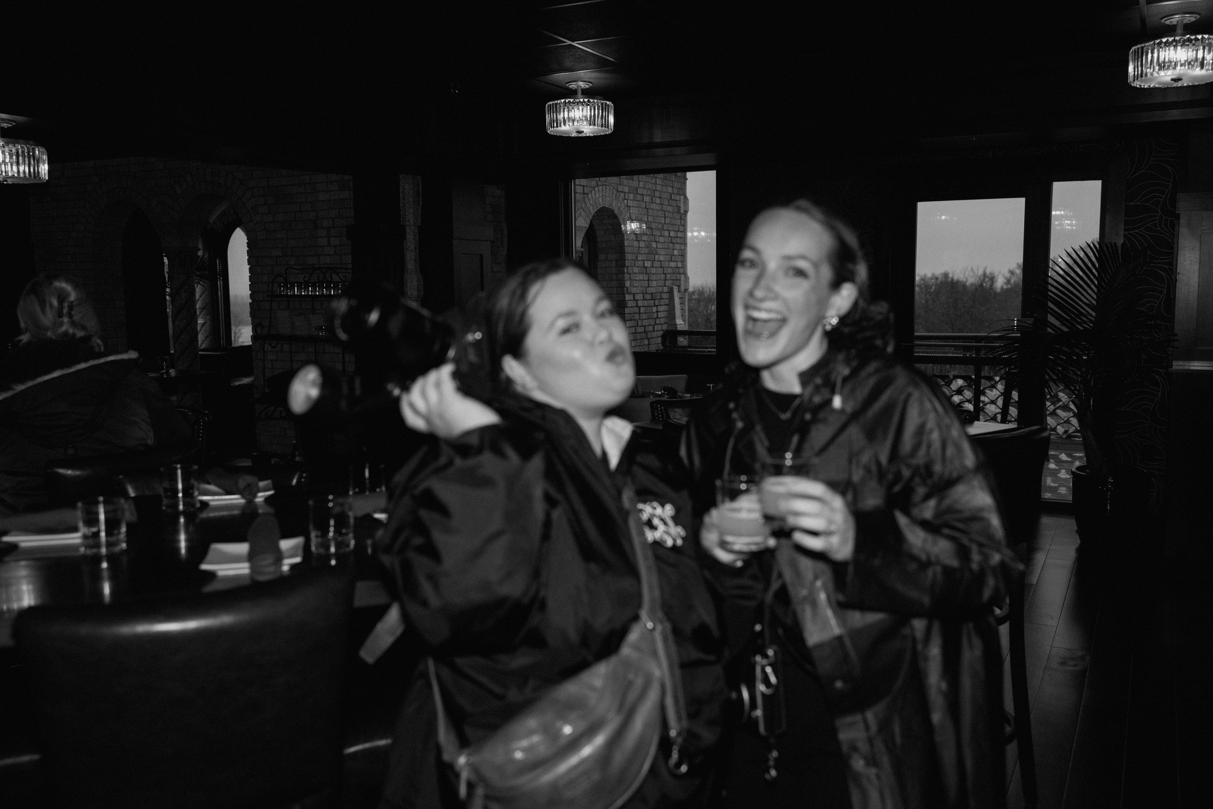 Two women celebrating with drinks in a dimly lit restaurant or bar, one holding a bottle and the other with a glass, smiling and making playful faces.