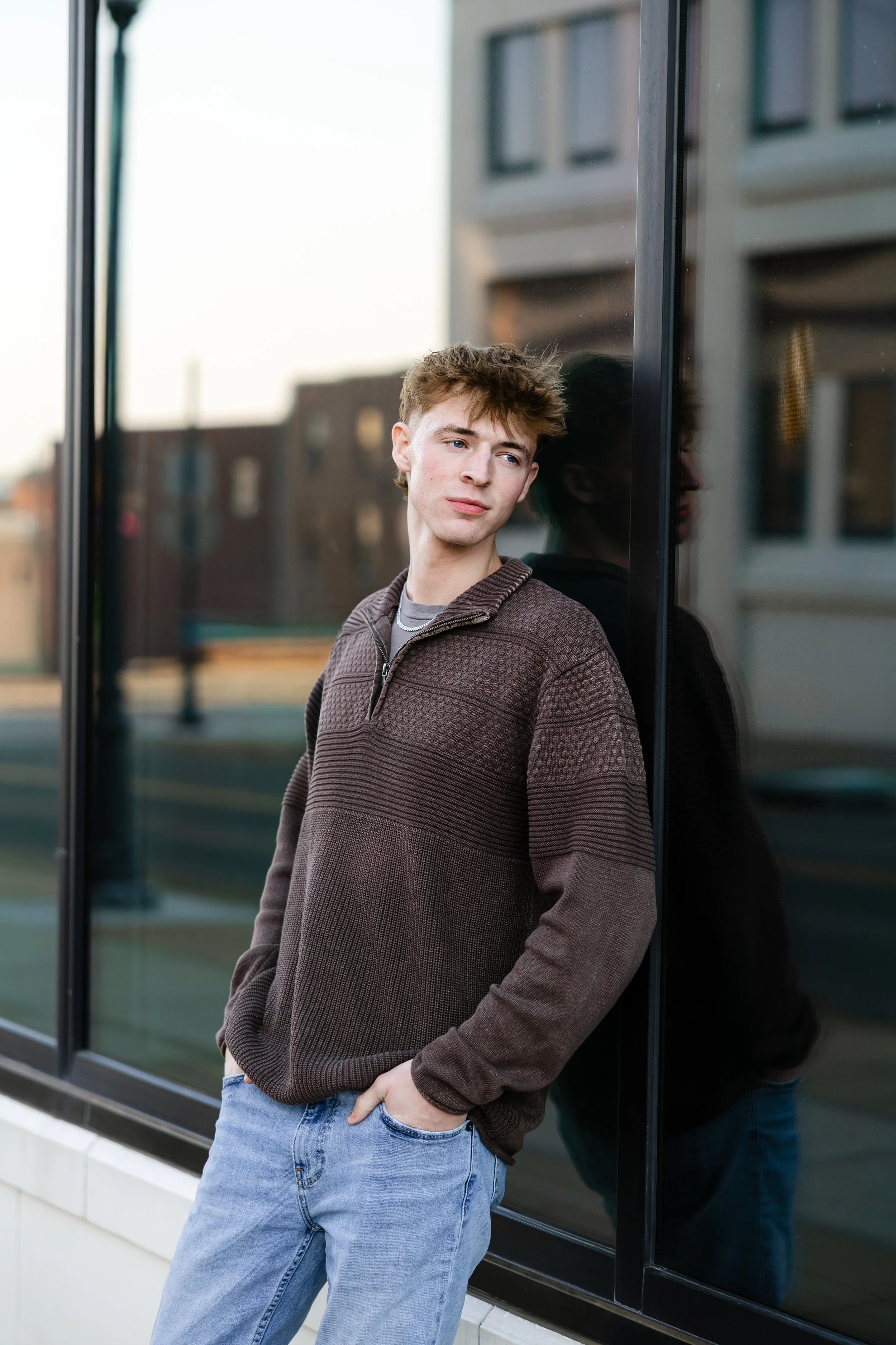 A young man with light brown hair, wearing a dark brown textured sweater and light blue jeans, leaning against a glass wall with a reflection of himself, in an urban environment.