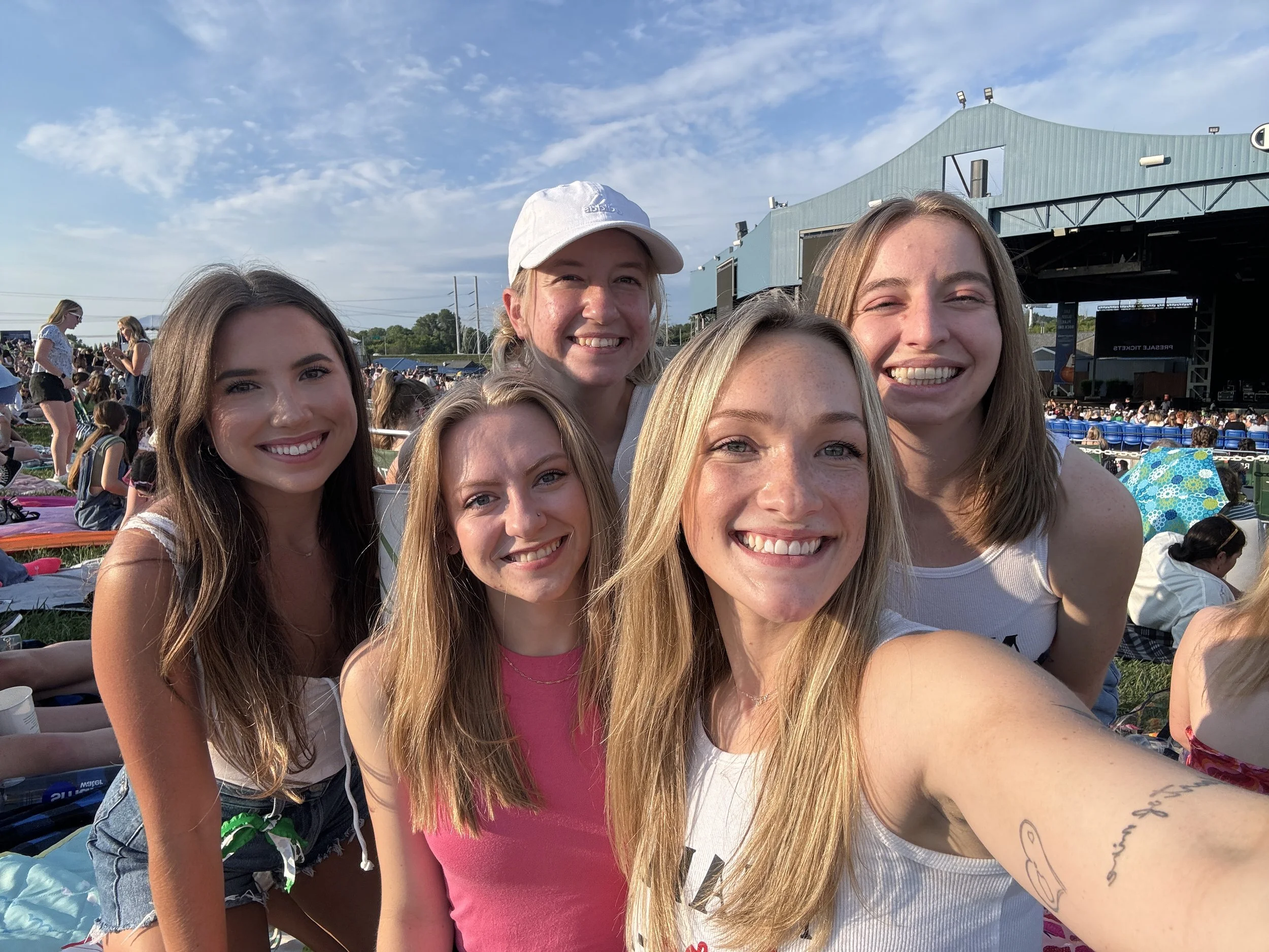 Group of five young women smiling, taking a selfie at an outdoor concert or event during daytime, with a stage and audience in the background.