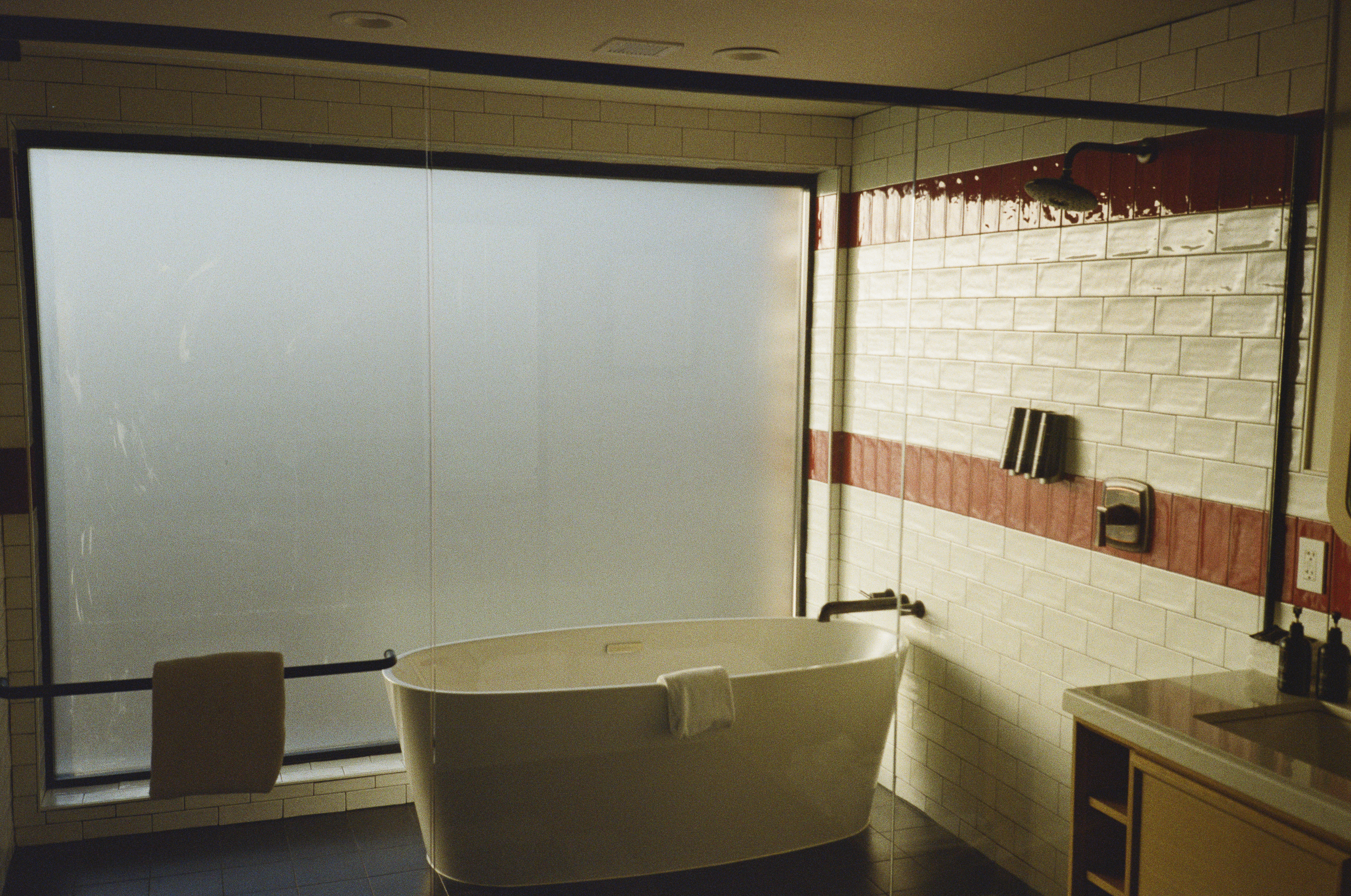 A bathroom features a standalone bathtub near a large frosted window, with a towel hanging over its edge. The wall has beige tiles with a red accent stripe, and a showerhead is visible. A sink with bottles is on a wooden cabinet.