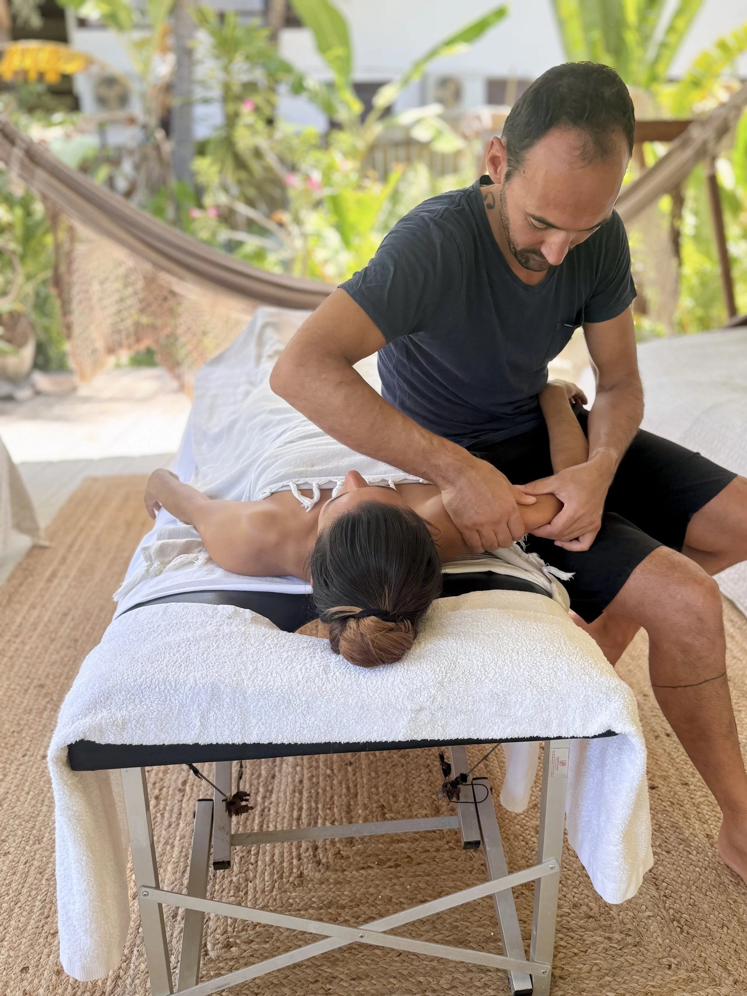 Un homme pratique une séance de massage ou de thérapie sur une femme allongée sur une table de massage dans un environnement naturel avec des plantes tropicales en arrière-plan.