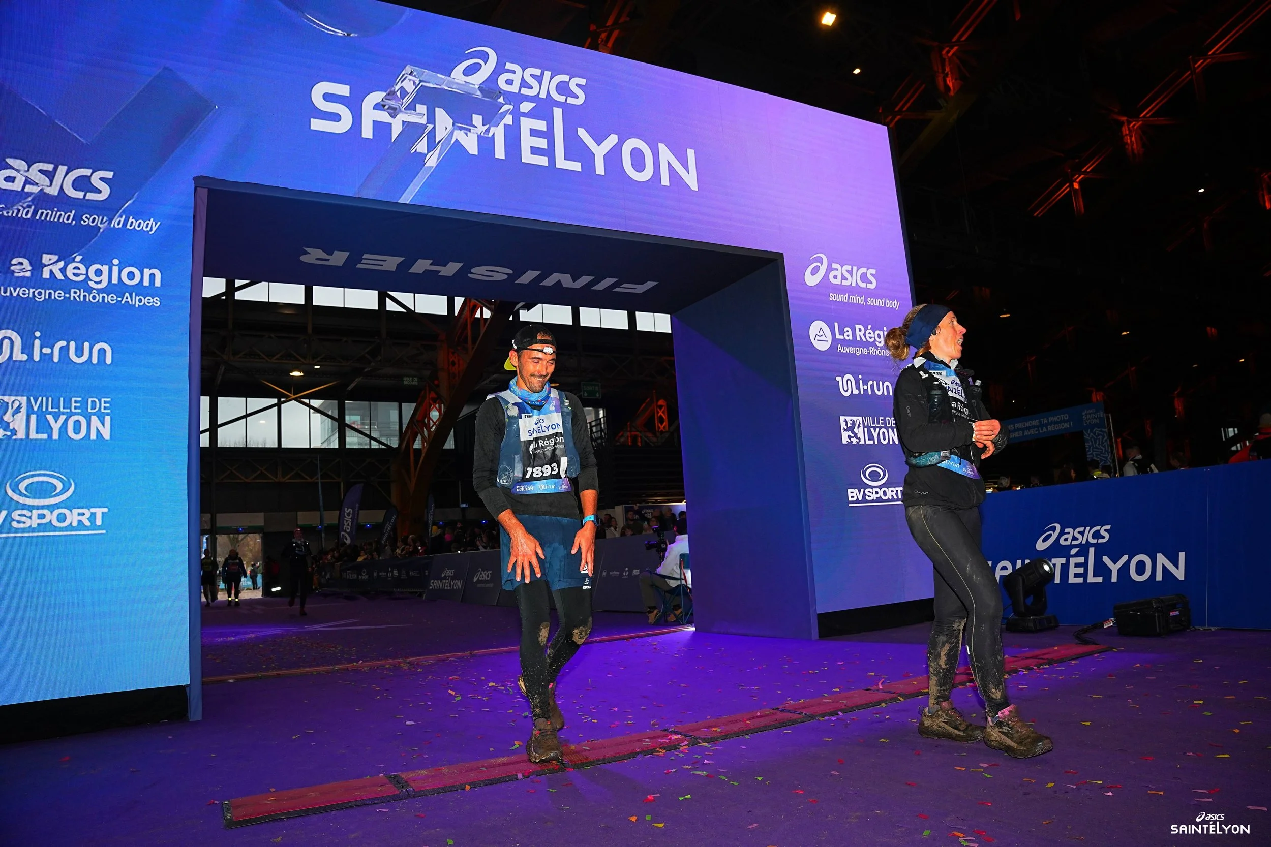 Deux personnes franchissent la ligne d'arrivée de la course SaintéLyon, toutes deux en tenue de course, avec des bibs portant le numéro 7893, dans une ambiance festive sous des lumières violettes et un grand écran en arrière-plan.