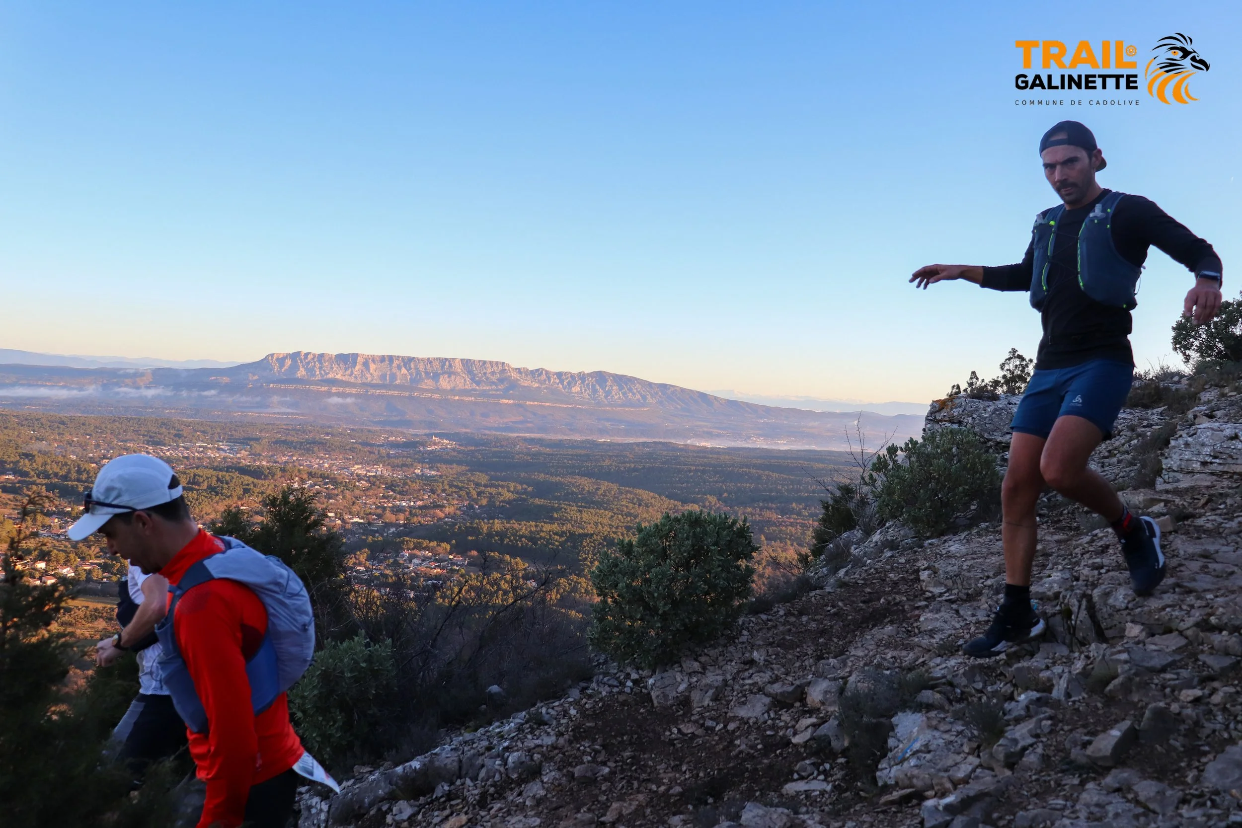 Trois personnes en trail running sur un sentier rocheux avec vue sur une plaine et des montagnes au loin, sous un ciel clair. Logo 'Trail Galinette' en haut à droite.