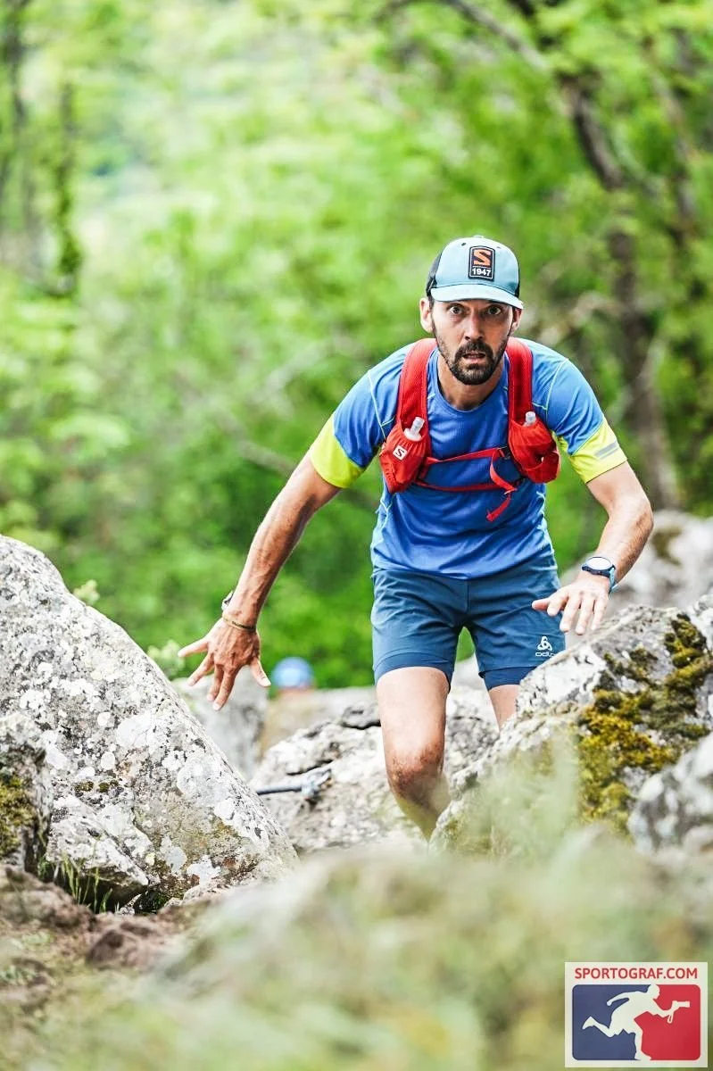 Un homme court dans une nature verte, entouré de rochers et d'arbres, probablement en train de faire de la course d'obstacles ou de la randonnée. Il porte une casquette, un t-shirt bleu avec des détails jaunes, un sac à dos rouge, une montre, et semb