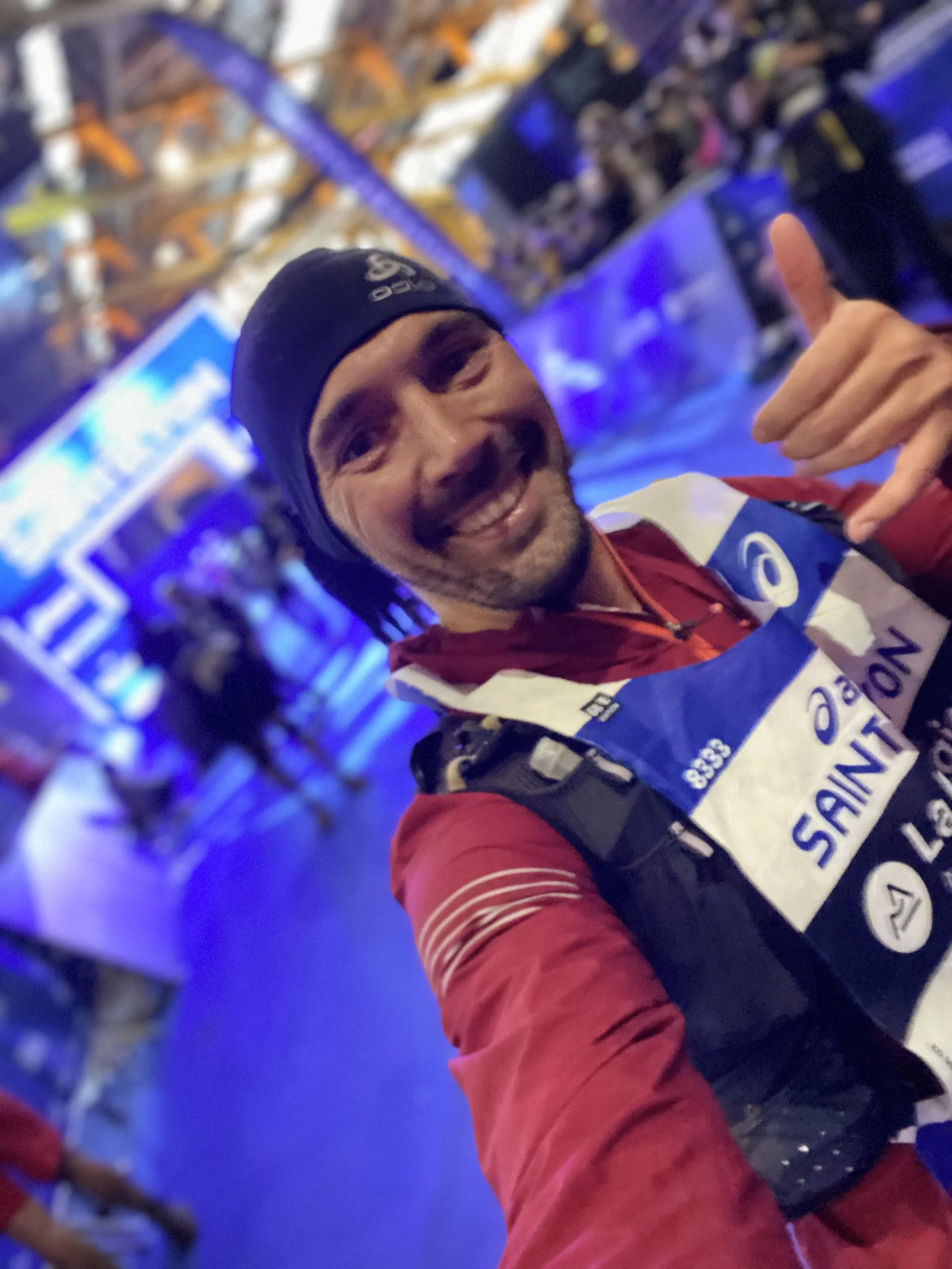 Homme souriant en tenue de course avec un dossard et un bonnet aux couleurs de Sainté, prenant un selfie dans une ambiance lumineuse bleue, avec des spectateurs et autres coureurs en arrière-plan.