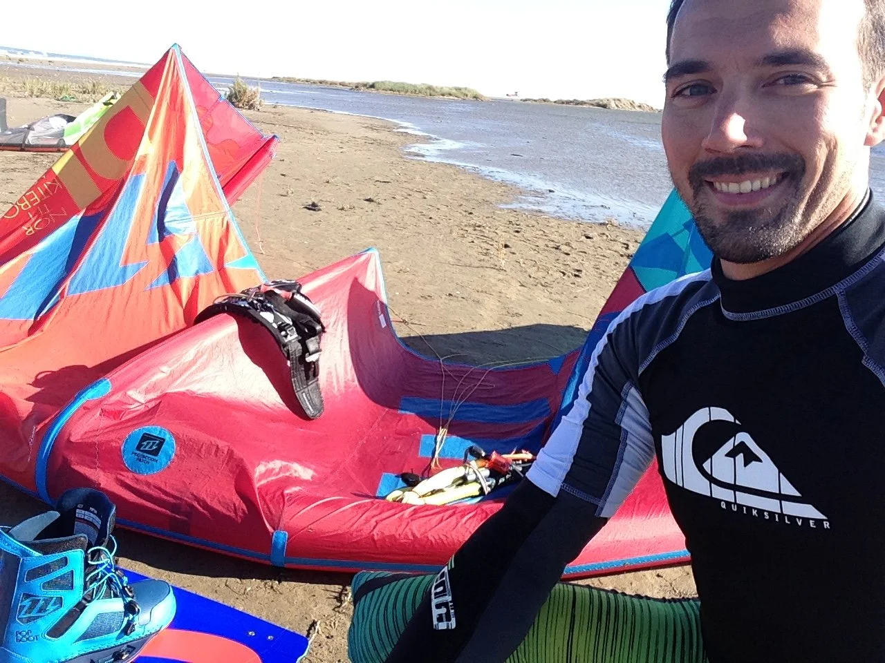 Un homme souriant portant un vêtement de sport Quiksilver, à côté d'une aile de kite sur la plage avec la mer en arrière-plan.