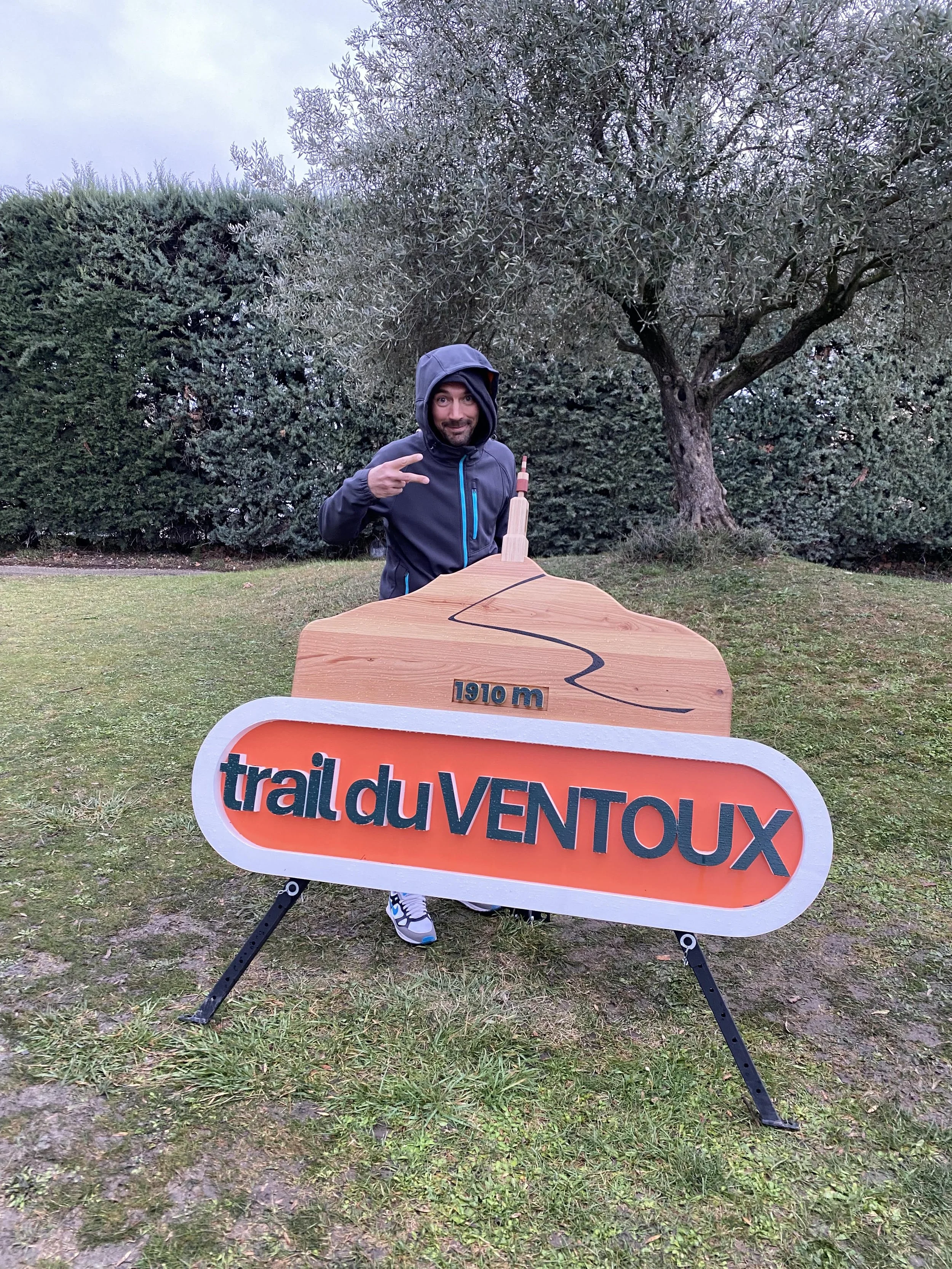 Un homme souriant, portant une veste à capuche, pointe vers un panneau indiquant le « trail du Ventoux » situé à 1910 mètres d'altitude, en extérieur avec des arbres en arrière-plan. Trail du Ventoux by Vieux Campeur 