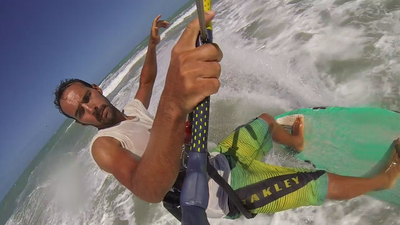 Homme faisant du kitesurf dans l'océan, portant un t-shirt blanc et un short vert, tenant la barre de kitesurf, avec une mer agitée et un ciel bleu en arrière-plan.