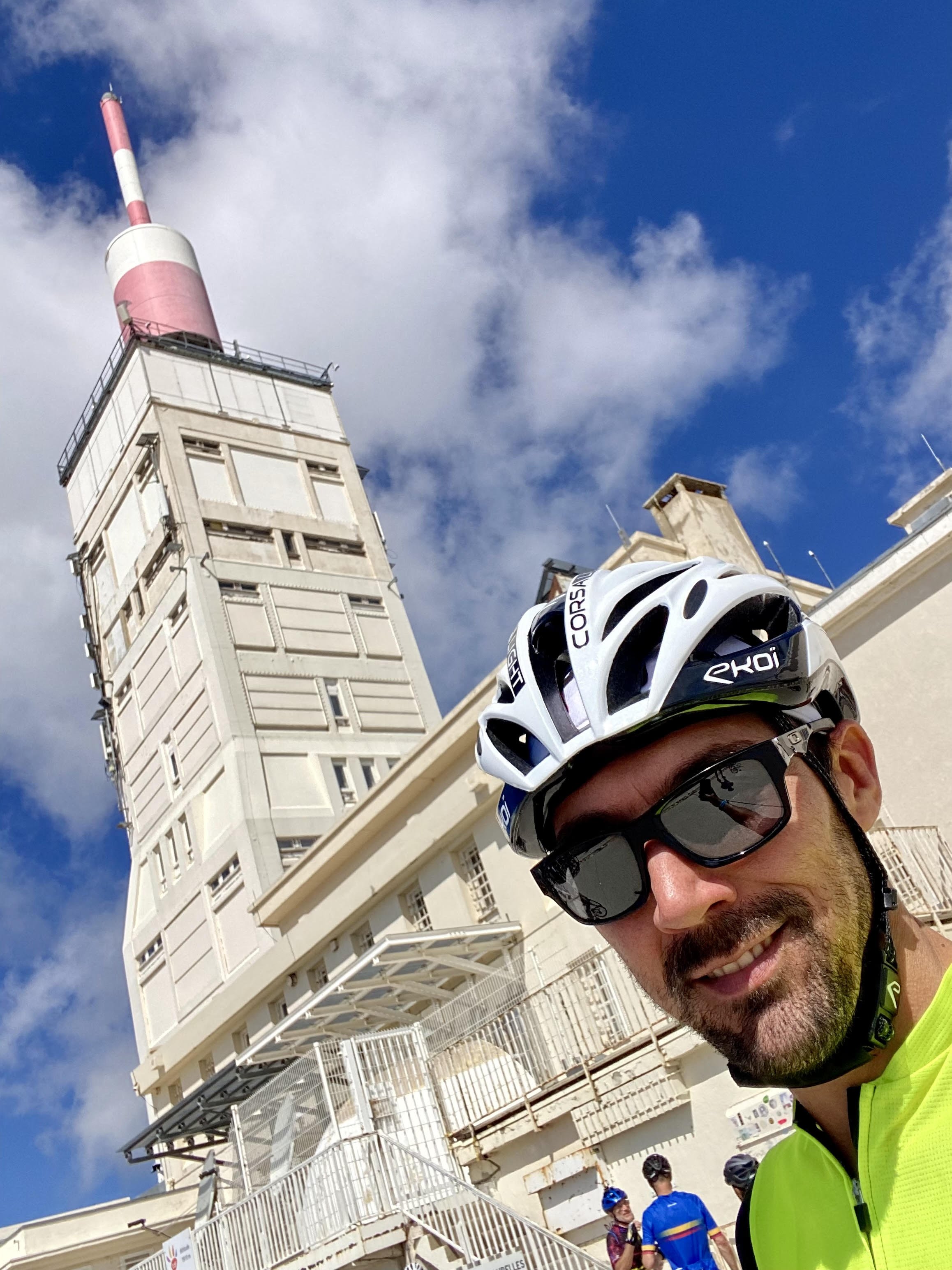 Homme souriant portant un casque de vélo et des lunettes de soleil, en premier plan, devant un bâtiment en hauteur avec un micro-ondes en haut, sous un ciel bleu avec quelques nuages. Mont Ventoux