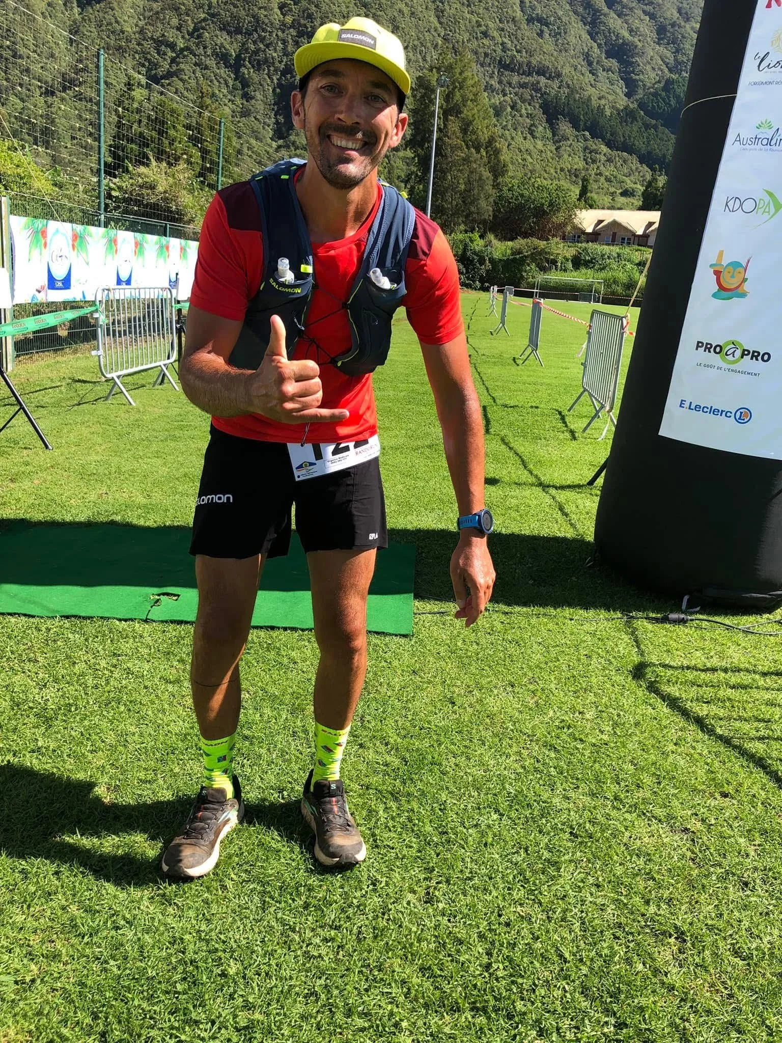 Un homme souriant, en tenue de course, se tient sur l'herbe d'un terrain en plein air, donnant un signe de la main. Il porte un short noir, un t-shirt rouge, des chaussures de course, une casquette jaune, et un gilet d'hydratation. Trail des Makes 