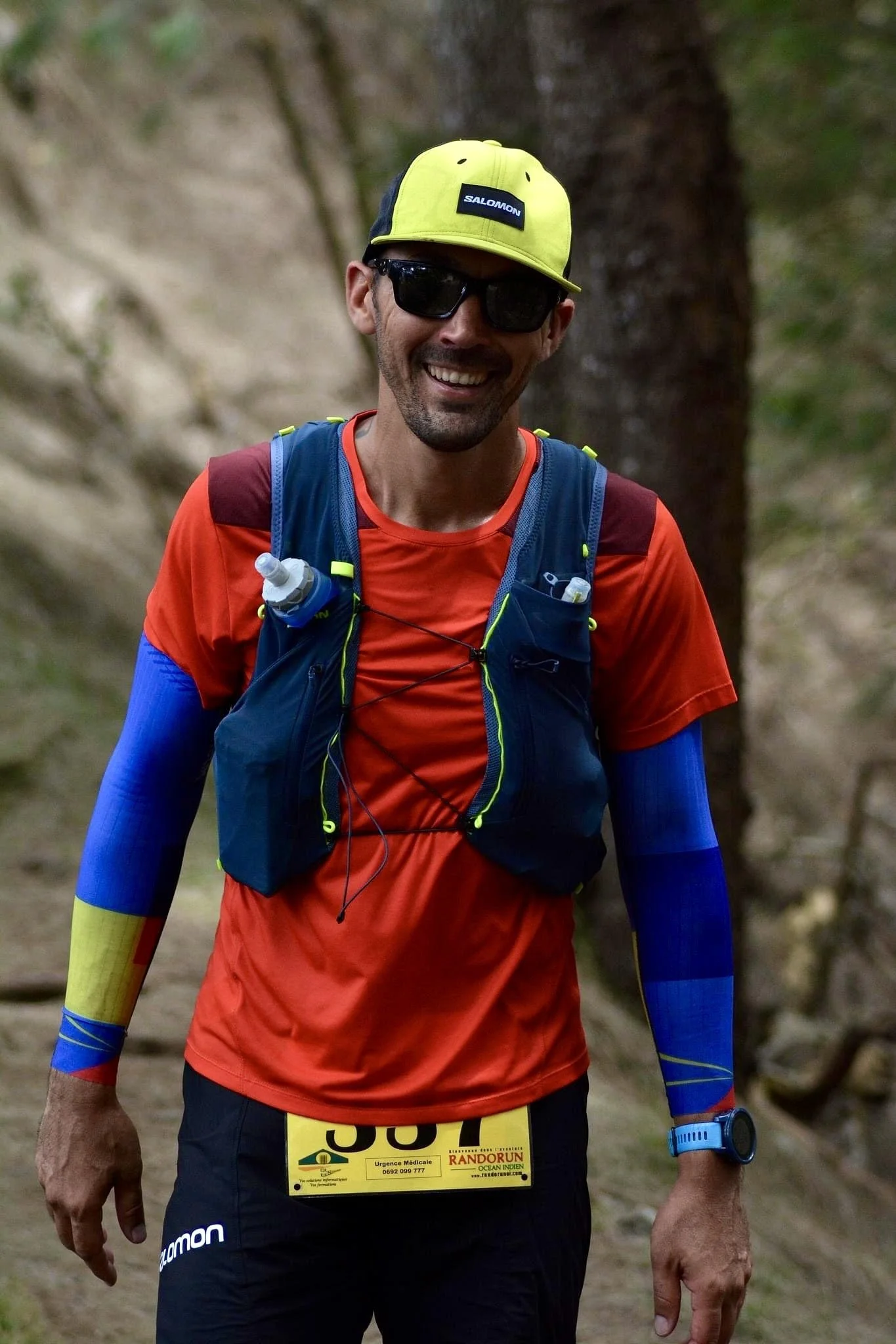 Coureur souriant portant un t-shirt rouge, un gilet de course, une casquette jaune, des lunettes de soleil, et un bracelet connecté, participant à une course en pleine nature. Mafate Trail Tour île de la Réunion