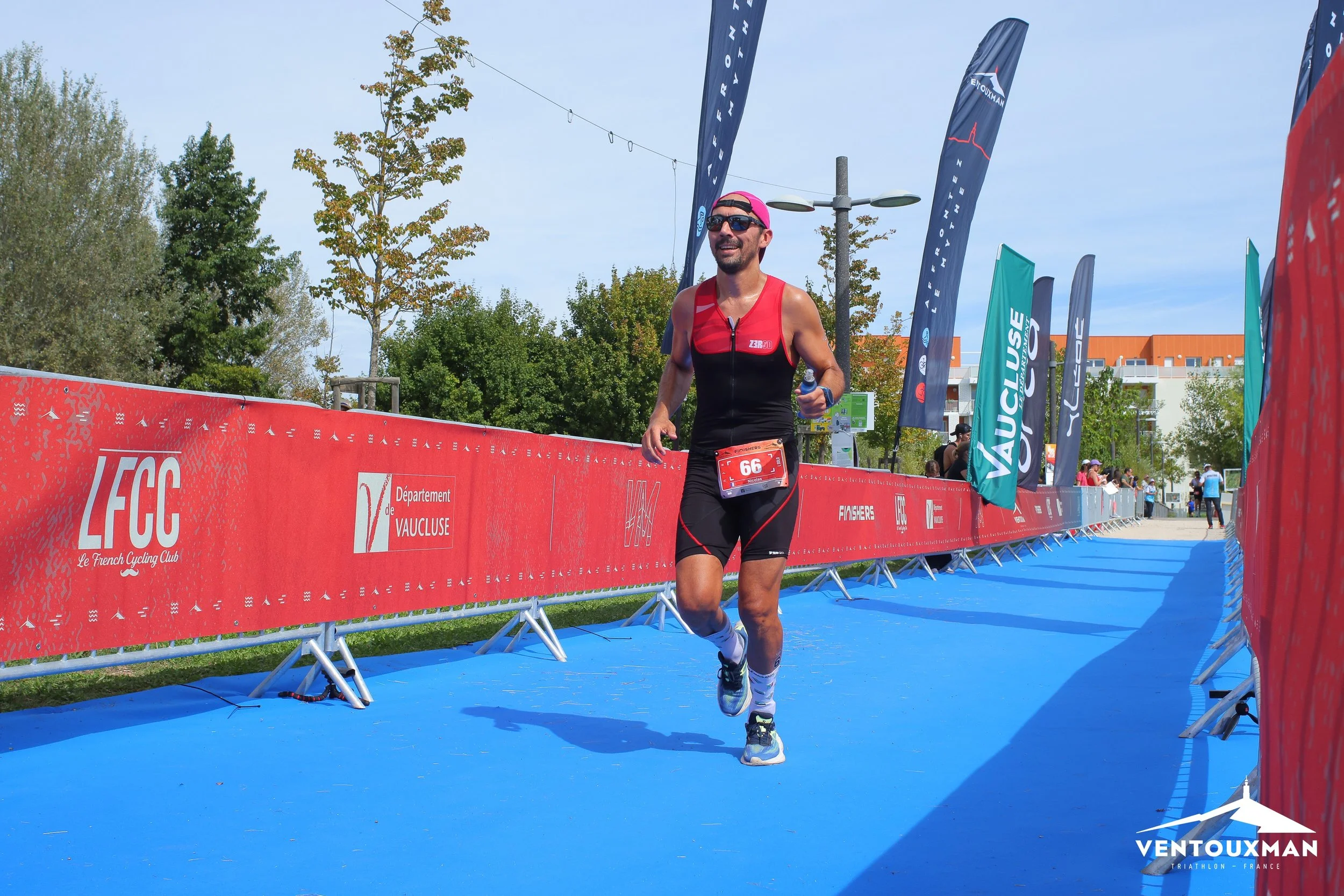 Un homme participe à une course de triathlon, il court sur un parcours bleu avec un dossard numéroté 66. Il porte une trifonction rouge et noir avec une casquette rose et des lunettes de soleil. L'arrière-plan montre des drapeaux et des spectateurs. 