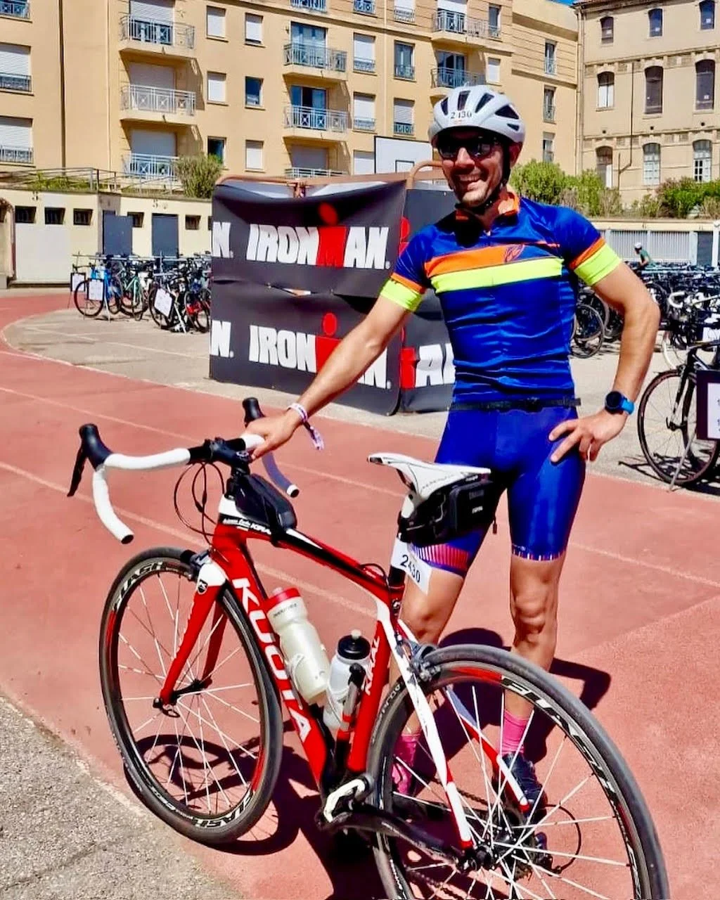 Un homme en tenue de cyclisme avec casque et lunettes, debout à côté de son vélo de route rouge, dans une zone de sport en plein air, avec des bâtiments en arrière-plan et un drapeau ou bannière Ironman. IronMAn 70.3 AIx en Provence