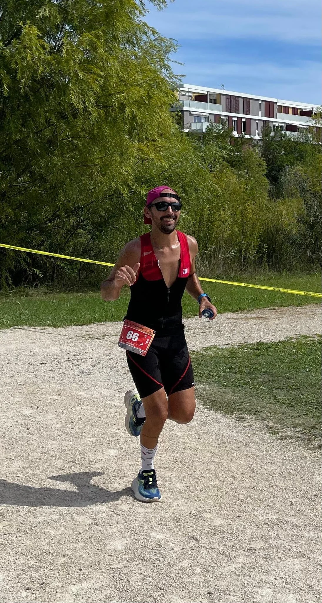 Un homme court lors d'une course en extérieur, portant un maillot noir et rouge, un numéro de course 66 attaché à sa taille, des lunettes de soleil, une casquette rose à l'envers, et tenant une bouteille d'eau. La scène est en plein air avec des arbr