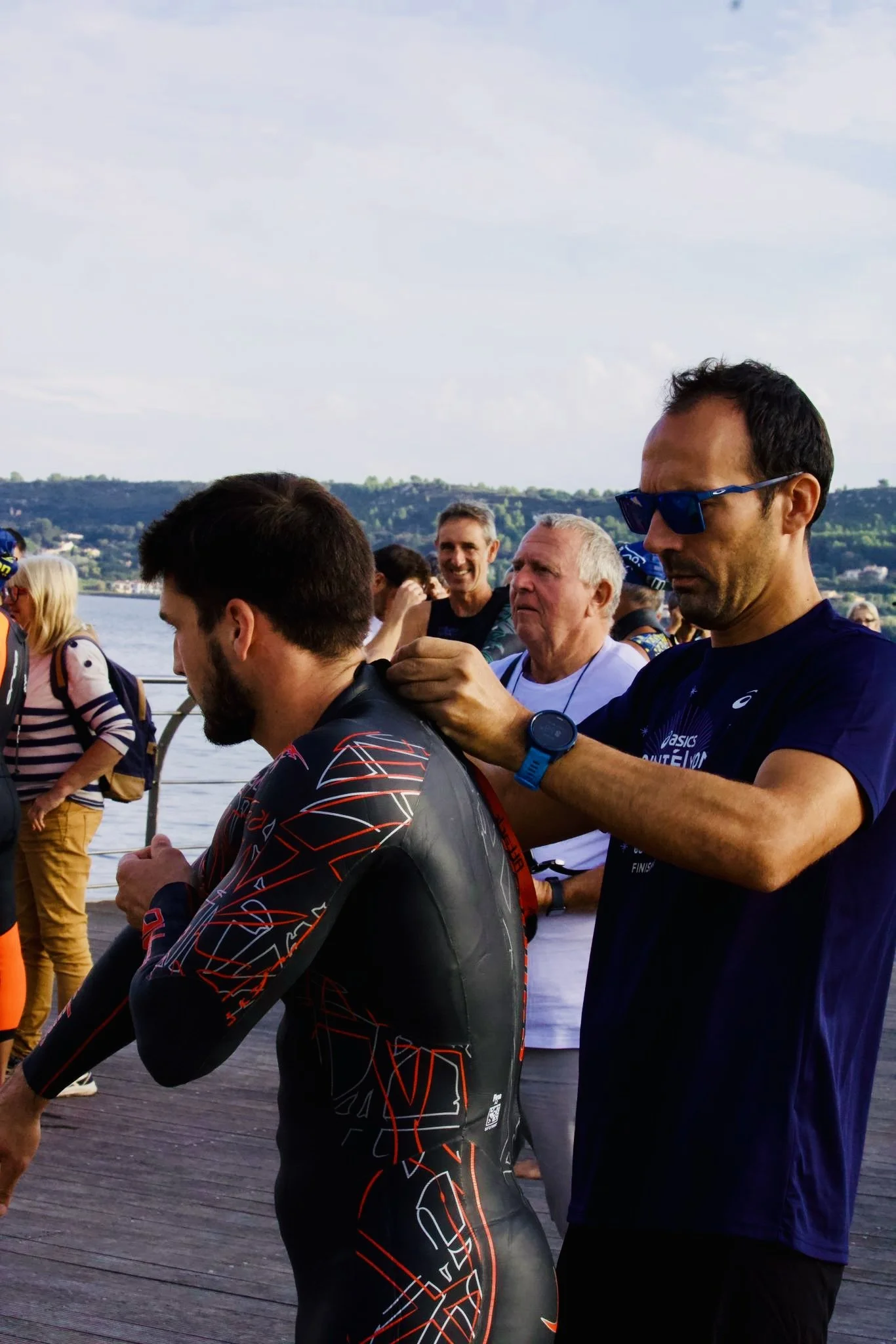 Un homme en combinaison de triathlon reçoit une cérémonie de fin de course par un autre homme portant des lunettes de soleil. D’autres personnes en arrière-plan regardent la scène, avec un paysage de rivière et de collines en arrière-plan.