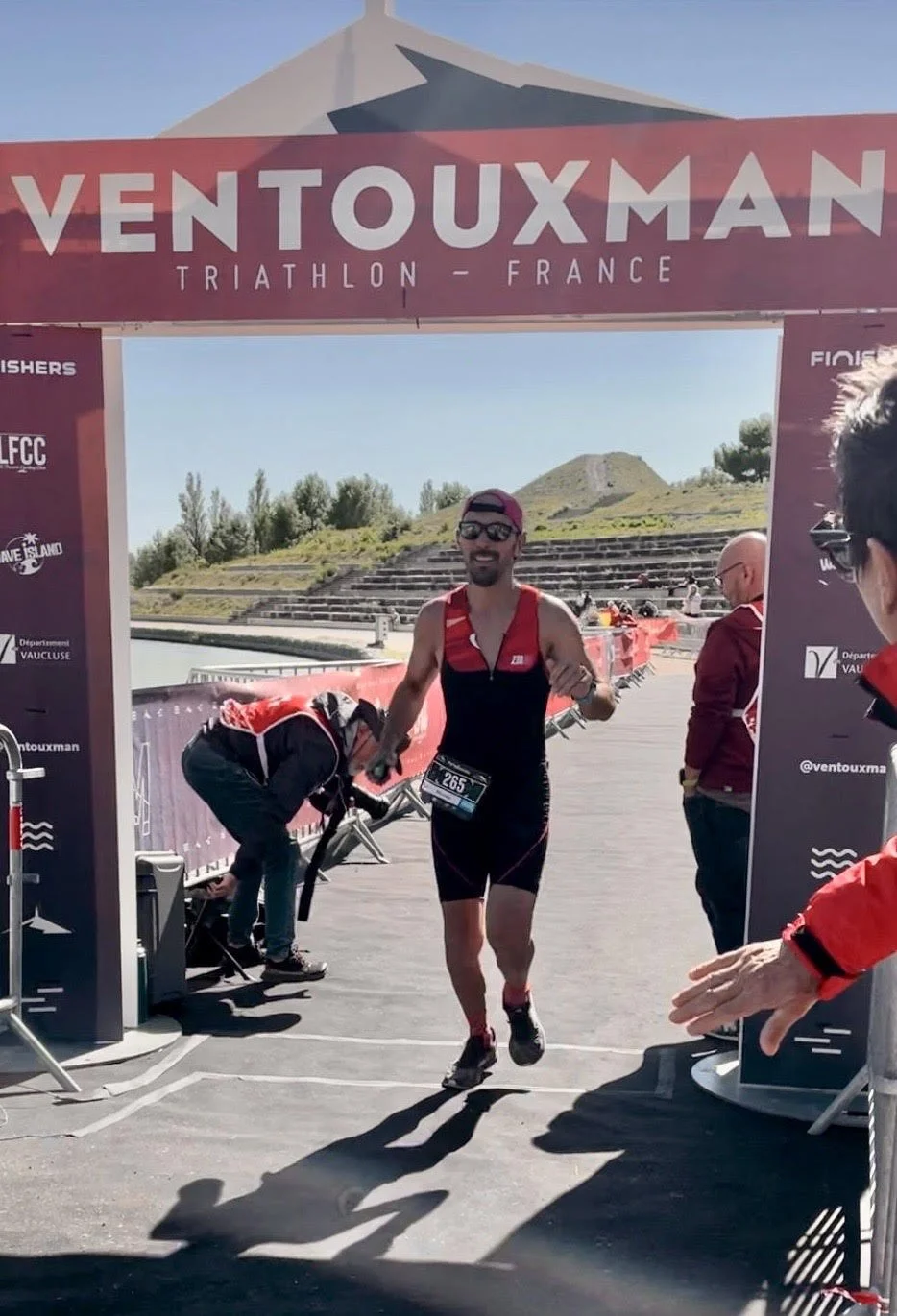Un homme en tenue de triathlon court à l'arrivée d'une course à Ventouxman en France, sous un panneau d'arrivée. L’arrière-plan montre des collines et un ciel clair. Il porte des lunettes de soleil, une casquette à l’envers, et un dossard avec le num