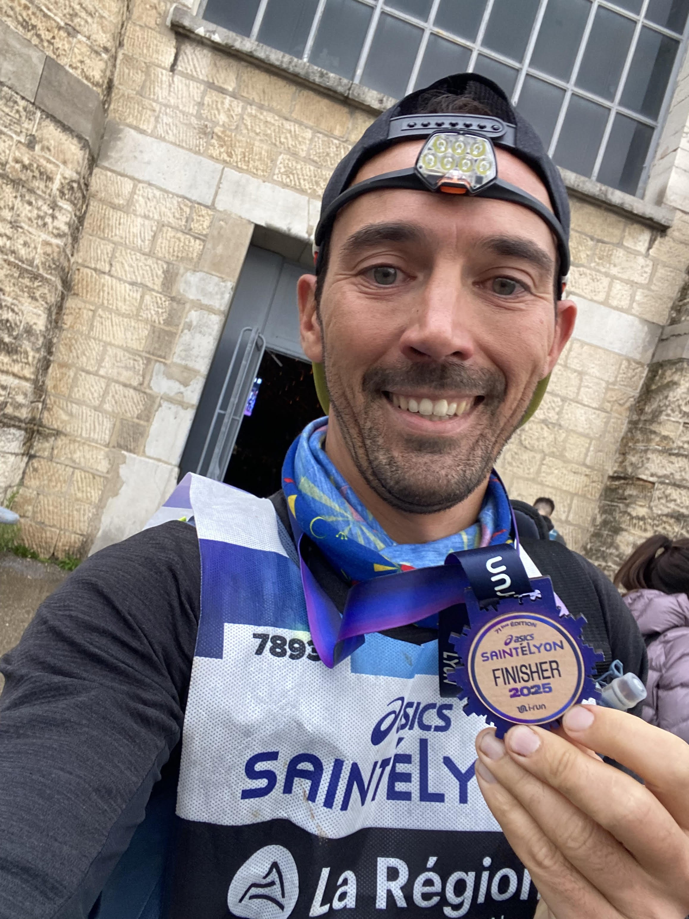 Un homme souriant portant une médaille de finisher de course à pied, portant une veste de course avec le nom 'Saintélyon 2025' et une lampe frontale, devant un bâtiment en pierre.