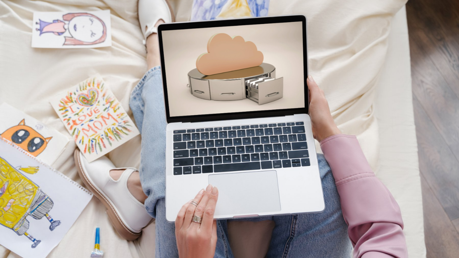 A person sitting on a bed, holding a laptop with a cloud storage icon on the screen. Around them are colorful drawings and a blue glue stick.