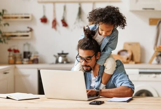 A man and a young girl are working together on a laptop in a kitchen. The girl is leaning over the man's shoulders, both focused on the screen.