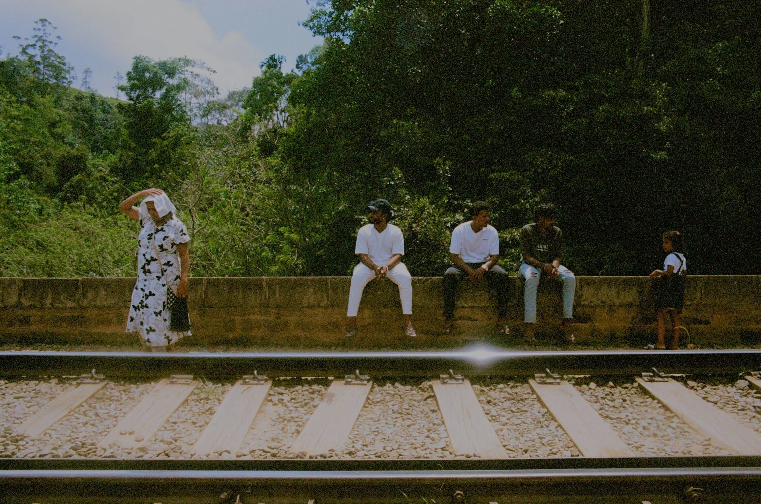 Four people sitting on a concrete barrier by railroad tracks with a woman standing nearby, set against a background of green trees and blue sky.