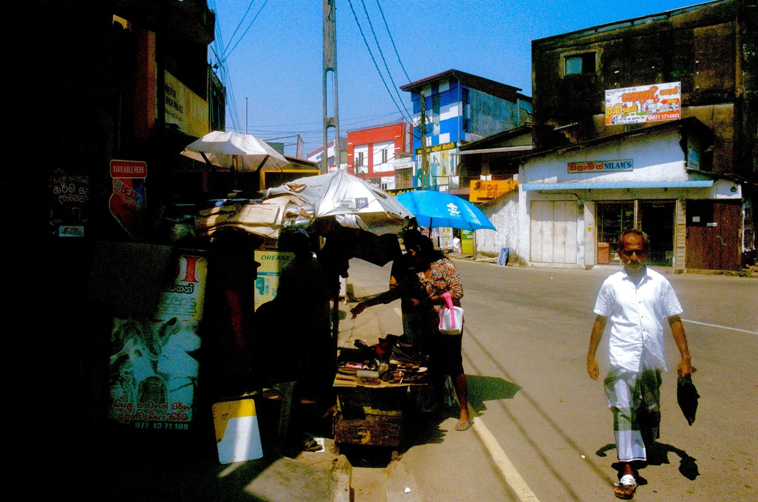 A street scene with two people shopping at a small roadside stall, one person under an umbrella and another walking on the sidewalk, with buildings and shops in the background on a sunny day.
