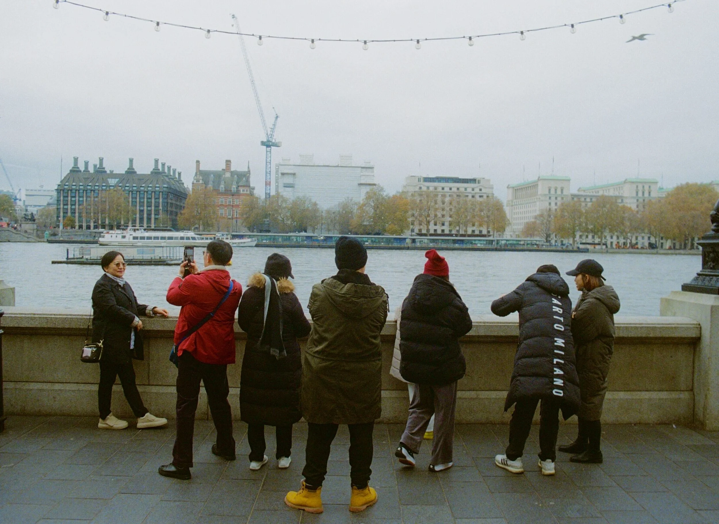 People standing and talking along a riverside promenade, with buildings and a boat visible across the river and string lights overhead.