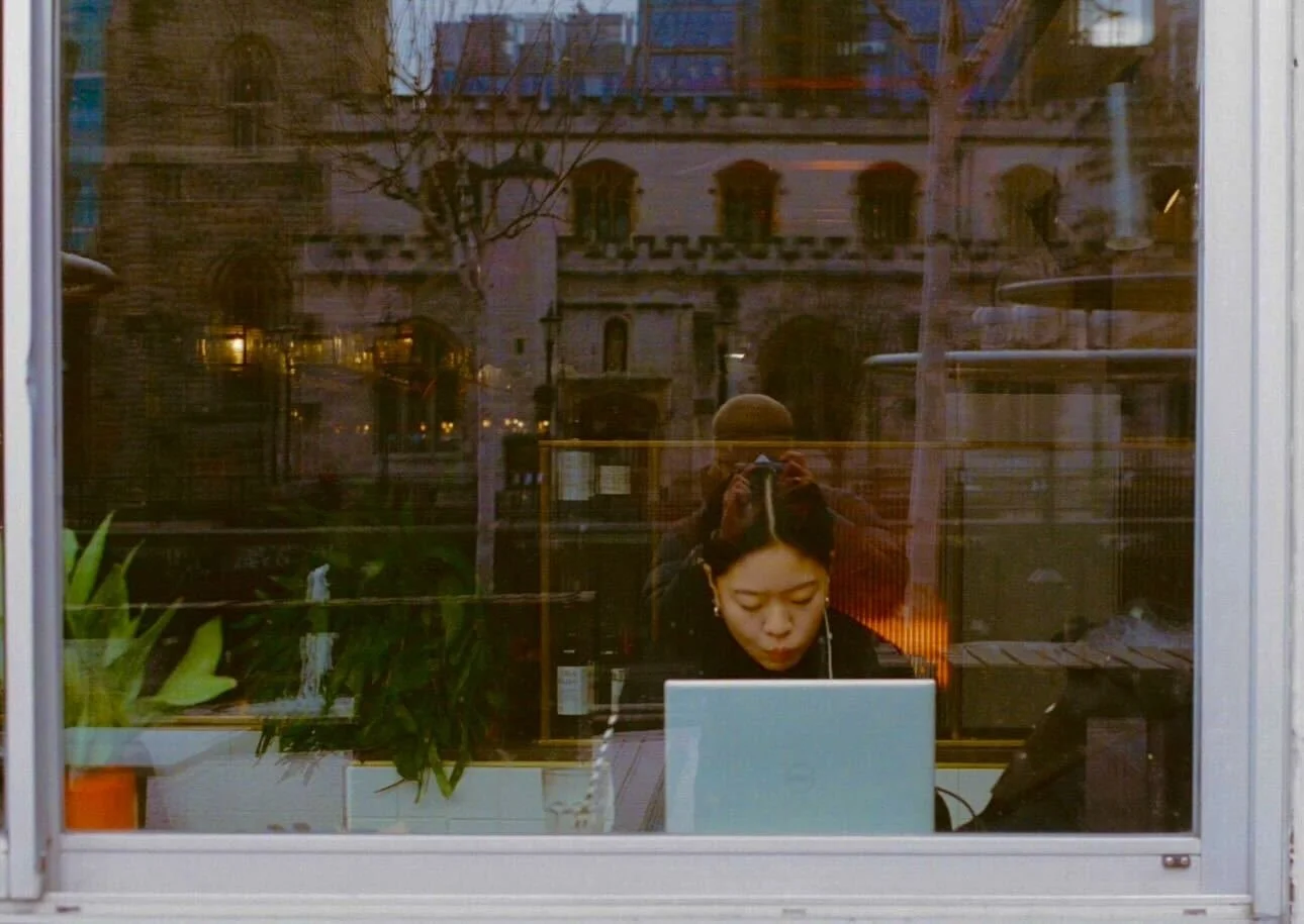 A person with dark hair working on a laptop, viewed through a window with a reflection of a person taking a photo, and an outdoor scene including buildings and trees.