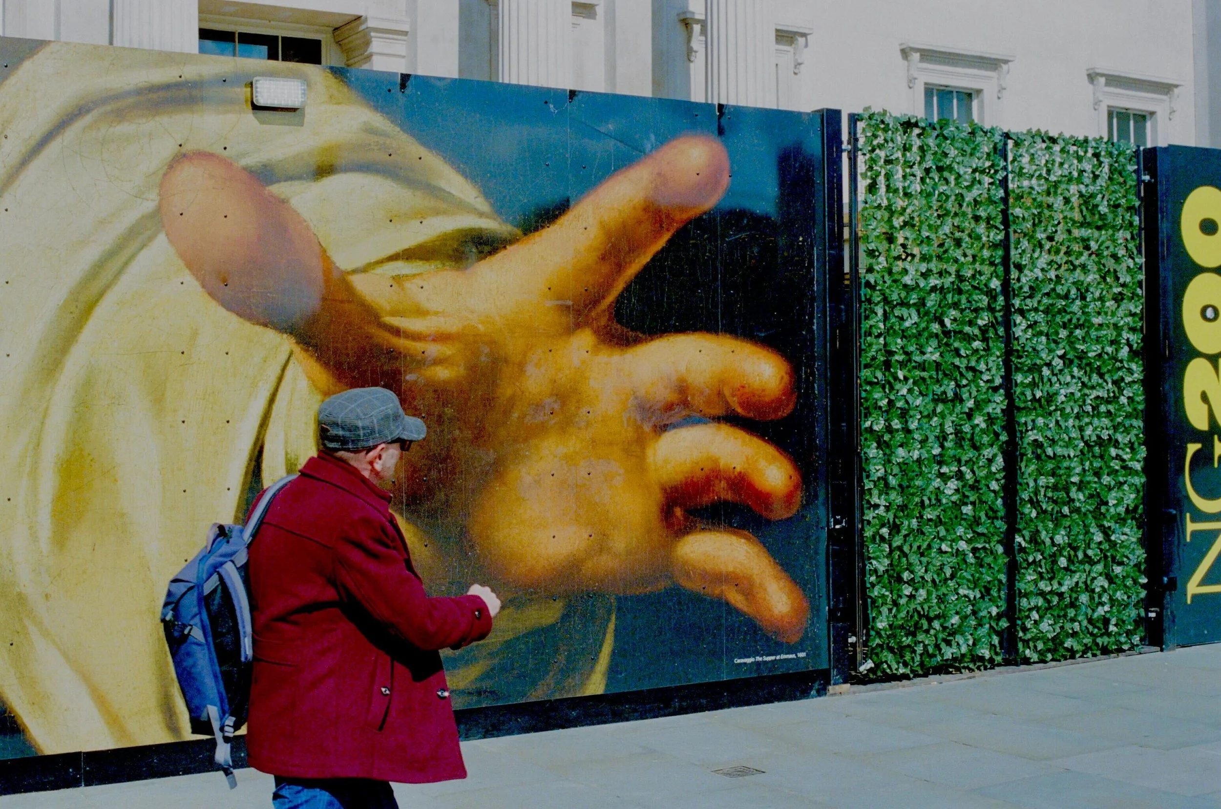 A man in a red coat and gray cap looking at a mural of a large hand. The mural is painted on a wall with greenery and buildings in the background.