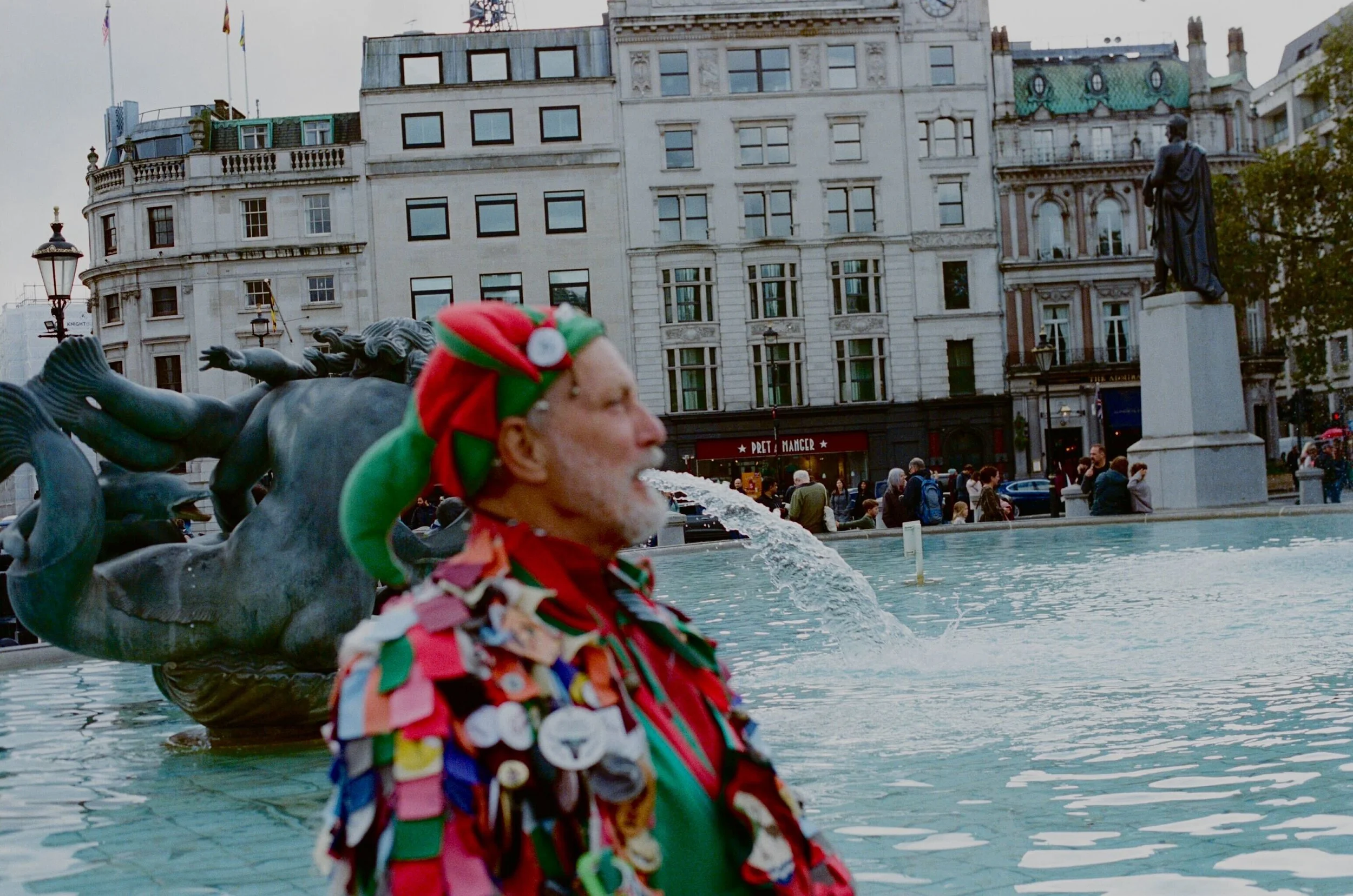 A man wearing a festive elf costume with buttons and badges standing near a fountain in Trafalgar Square, London, with white buildings and statue in the background.