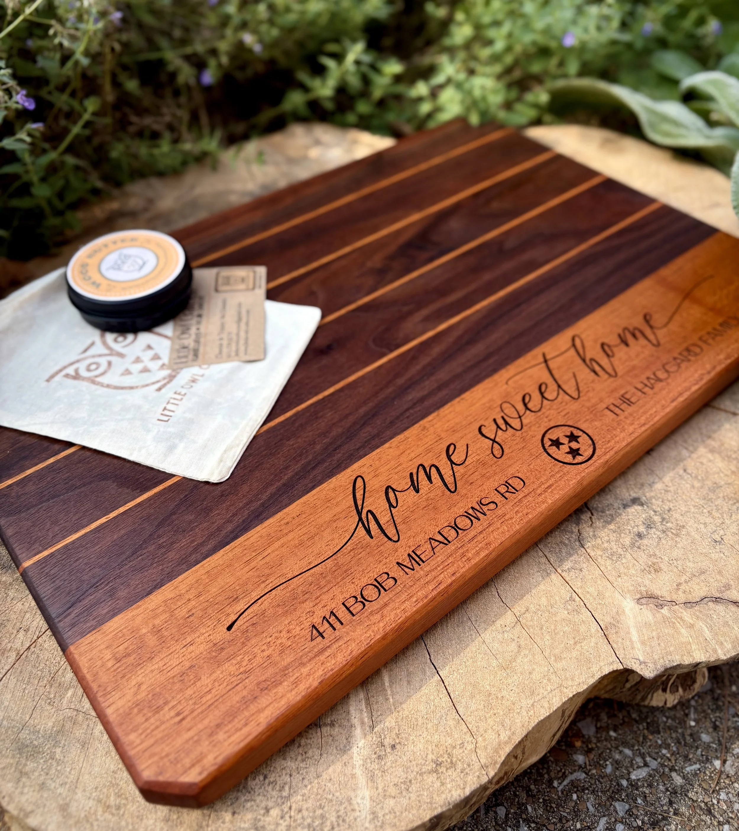 A wooden cutting board with the engraved phrase 'home sweet home' and an address '411 Bob McAdoms Rd' with a Tennessee state star logo, placed on a large piece of wood with outdoor greenery in the background.