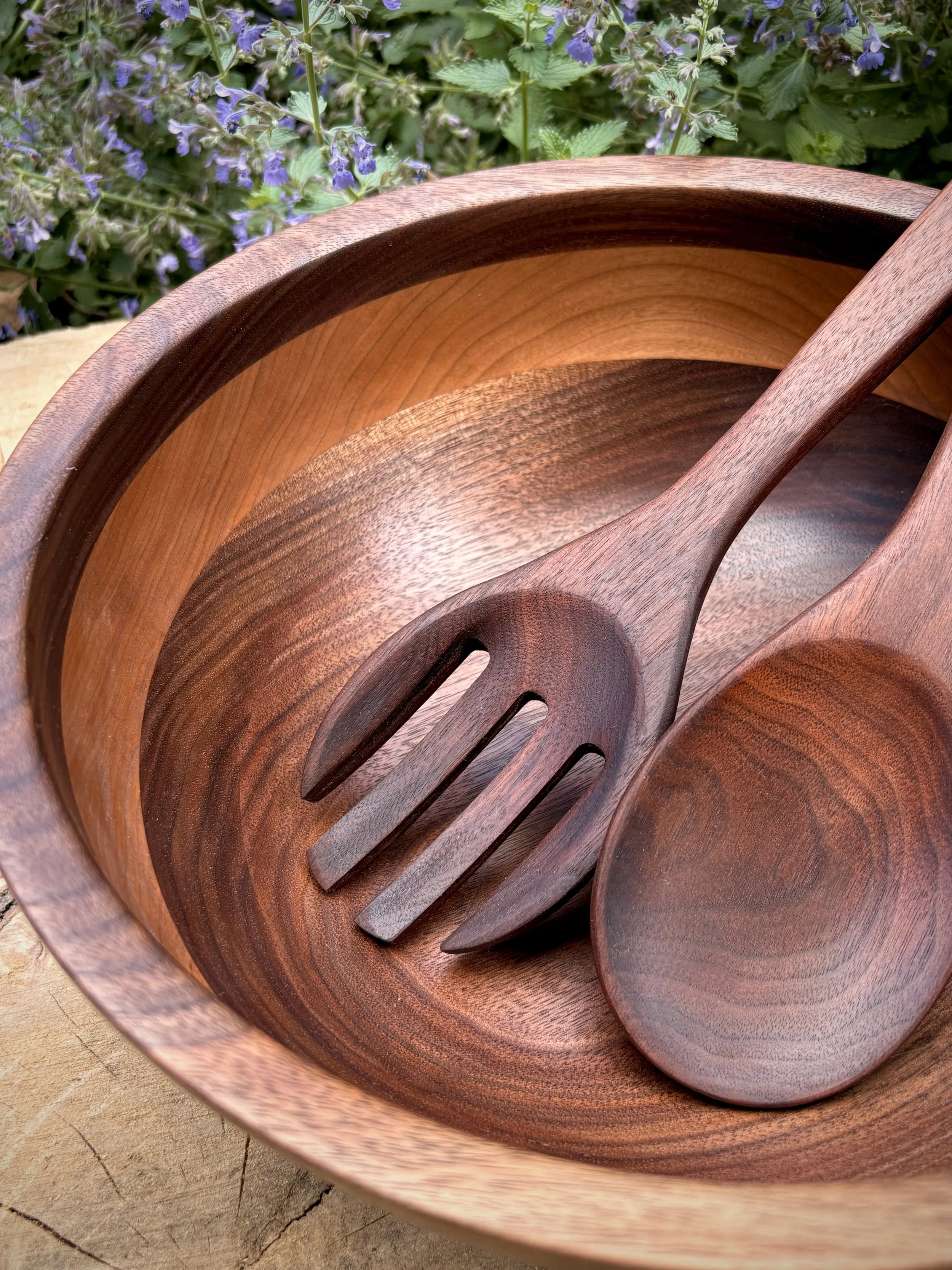 Close-up of a wooden bowl, spoon, and fork set placed inside a larger wooden bowl, with purple flowers and green leaves in the background.