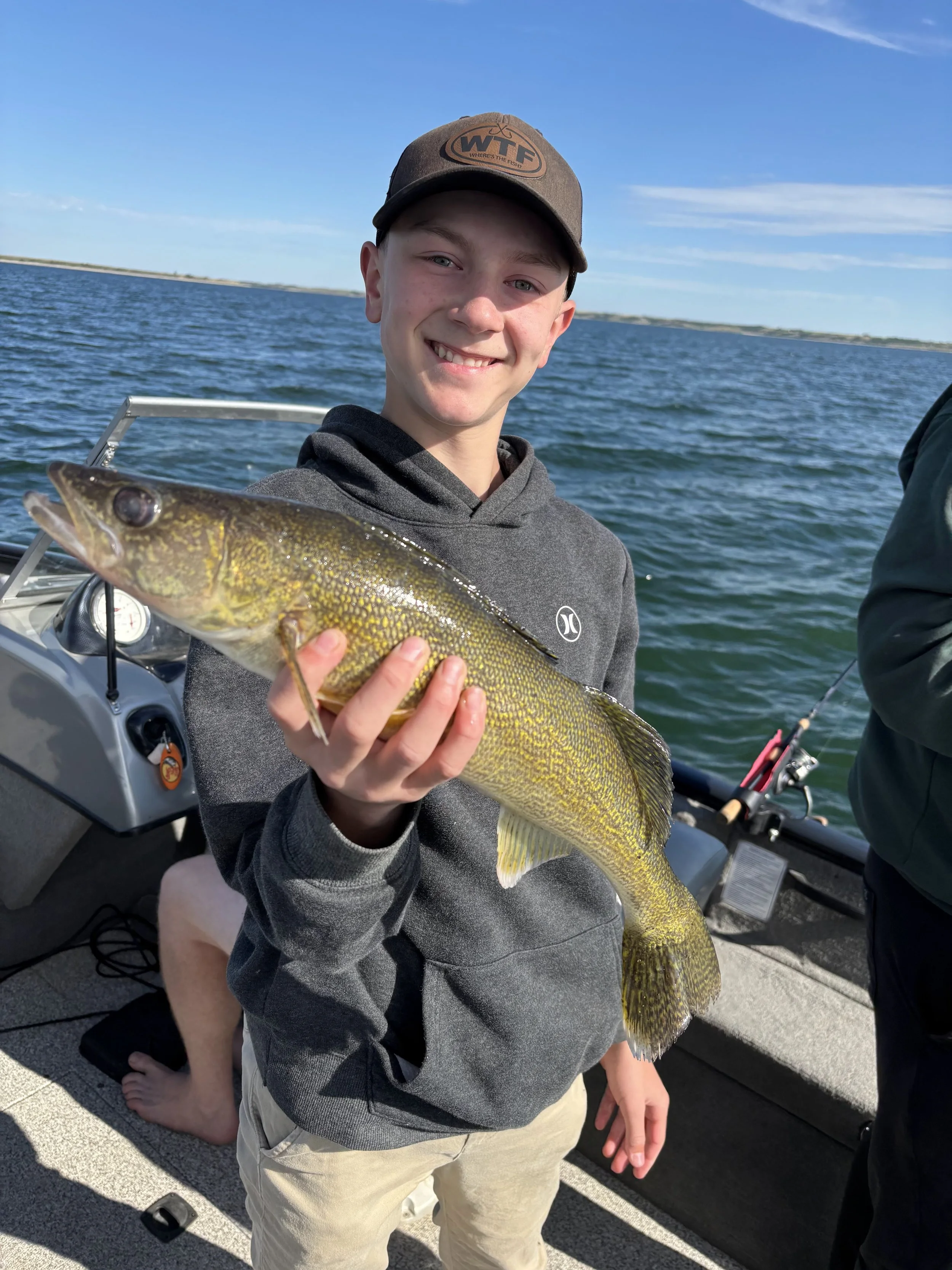 A young boy smiling on a boat holding a large fish, with water and a blue sky in the background.