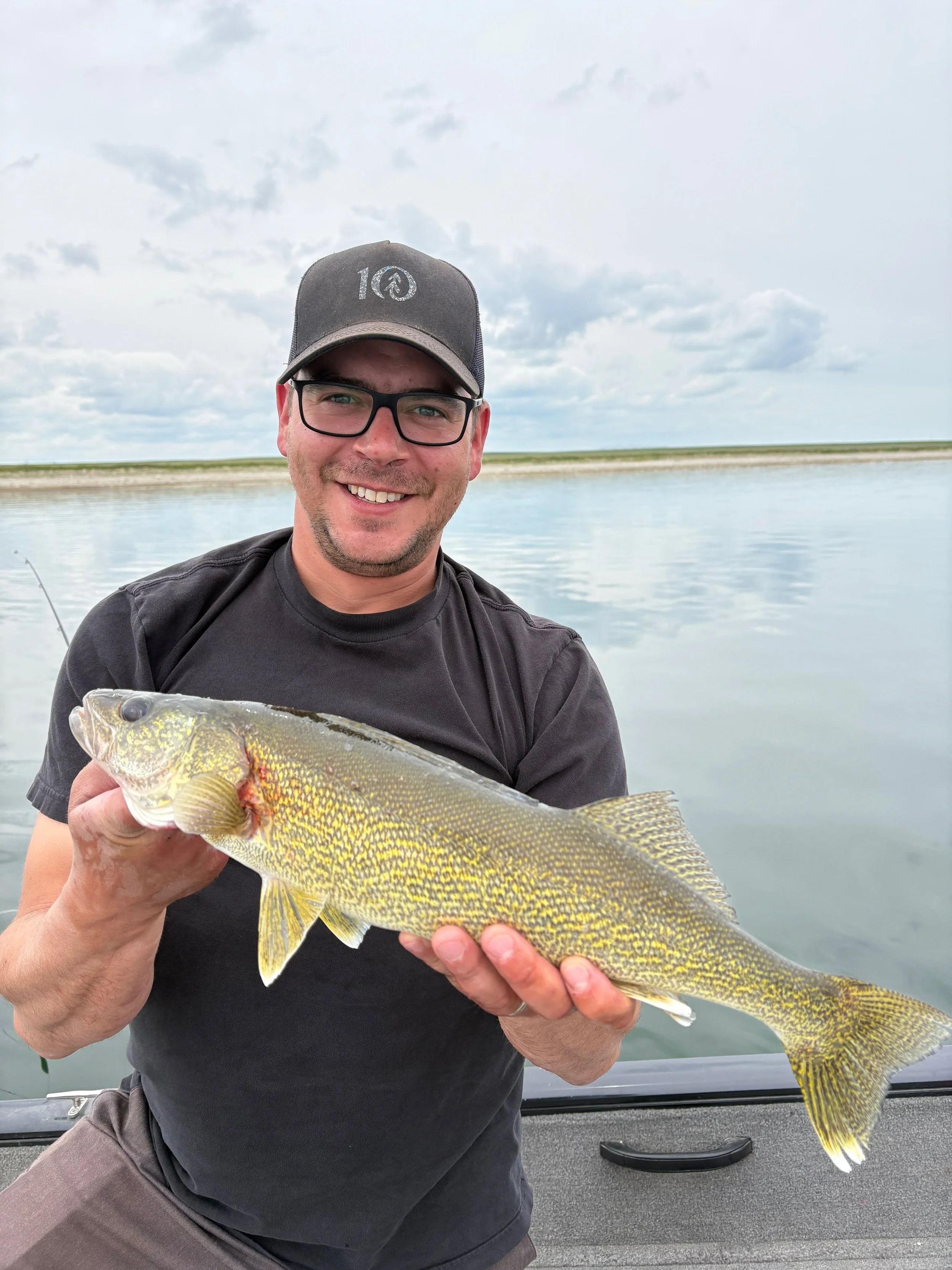 Man wearing glasses and a black baseball cap smiling while holding a large fish near a body of water with cloudy sky.
