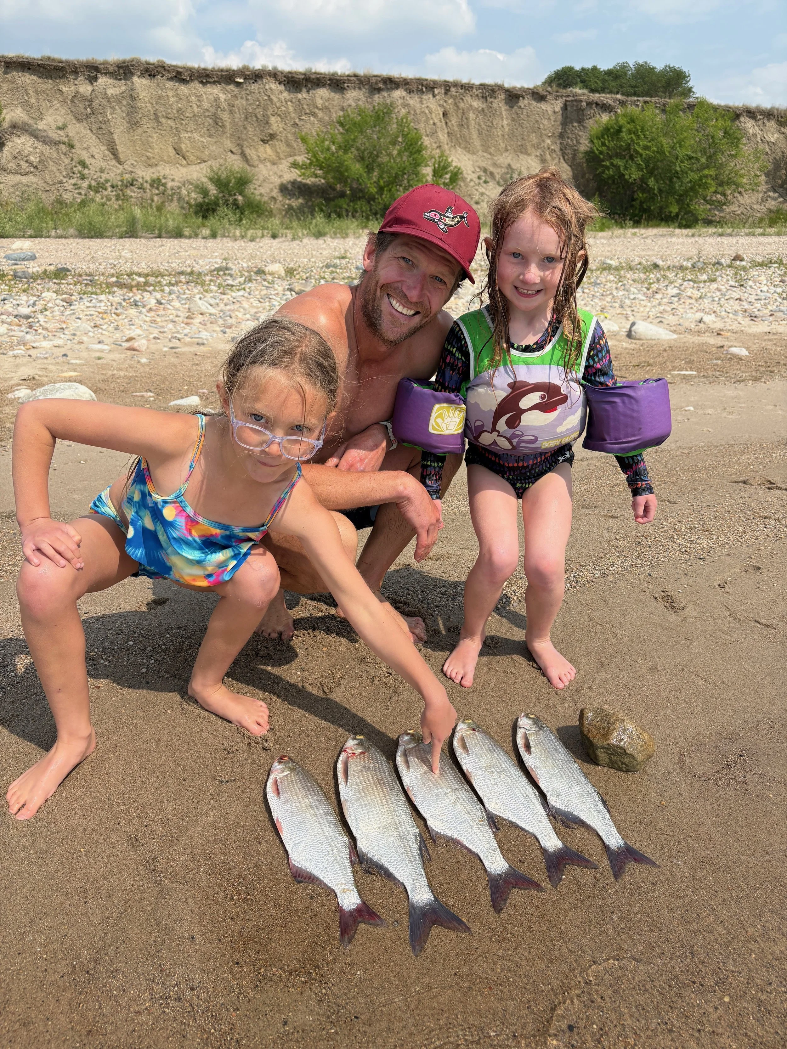 A man and two young girls on a sandy beach, showing off their caught fish, with a cliff and green trees in the background.
