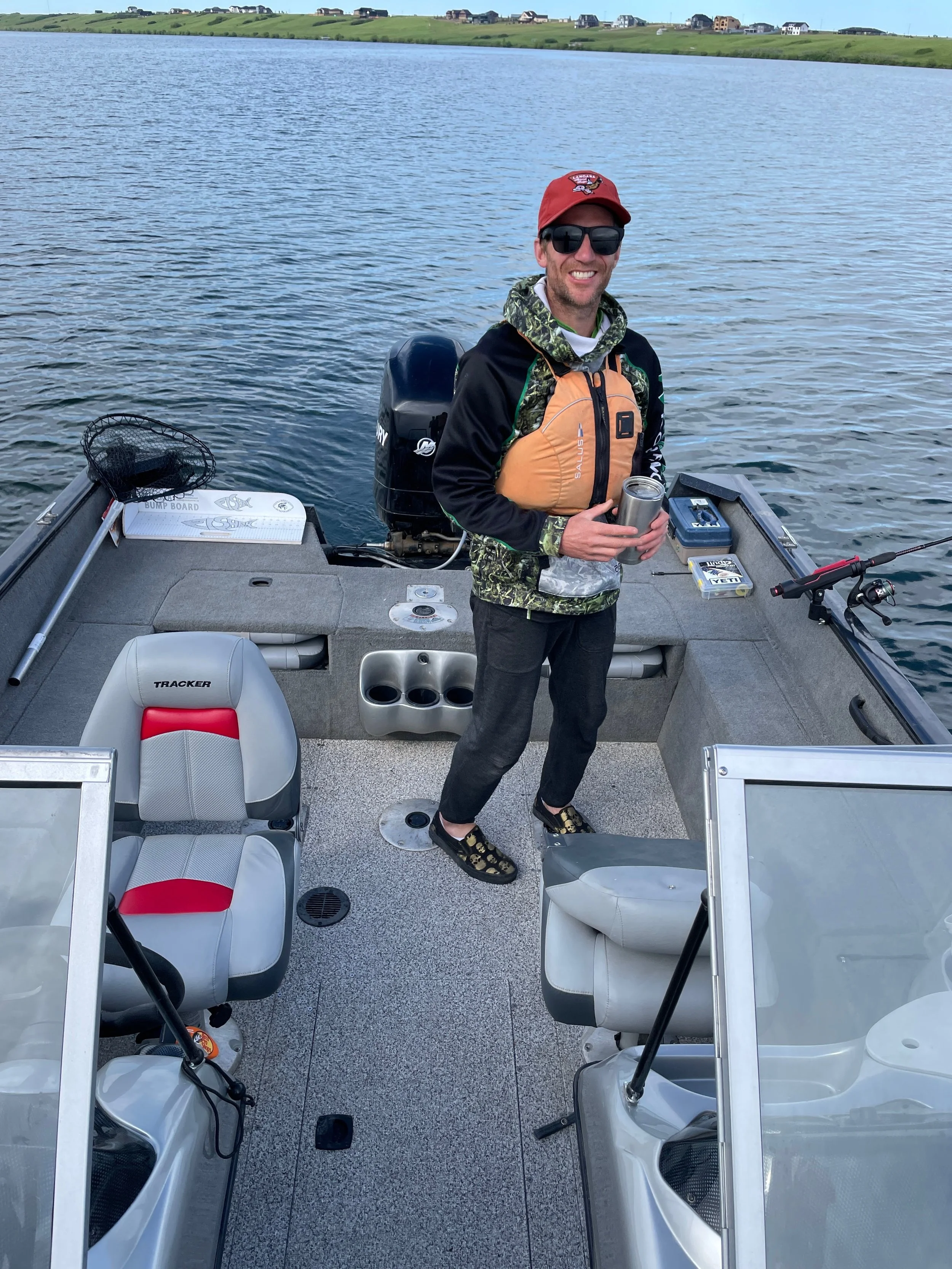 A man standing on a boat holding a tumbler, wearing sunglasses, a red cap, a camouflage hoodie, an orange life jacket, and sandals, with a lake and houses visible in the background.
