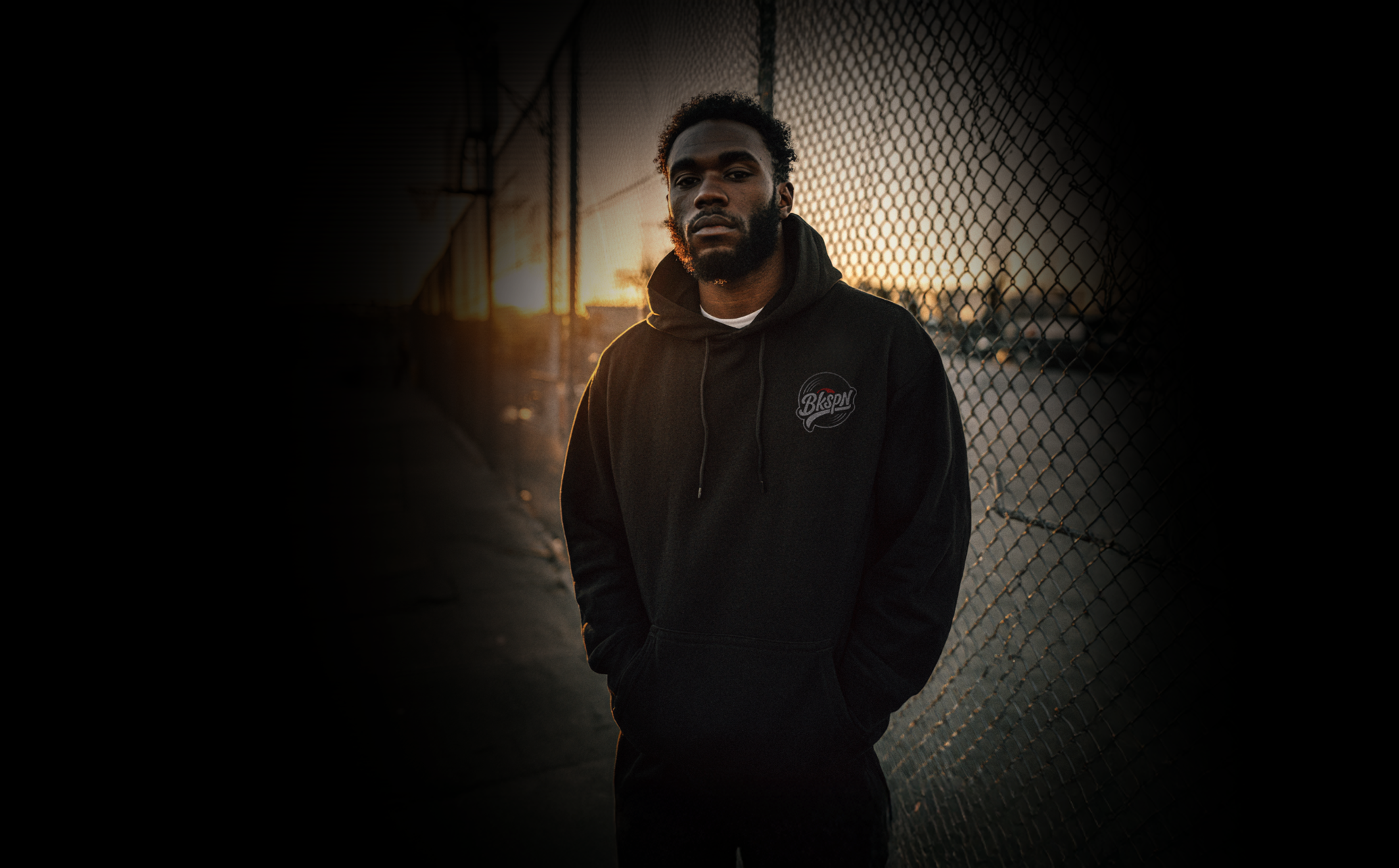 A young man standing by a chain-link fence at sunset, wearing a black hoodie with a BKSPN logo on it.