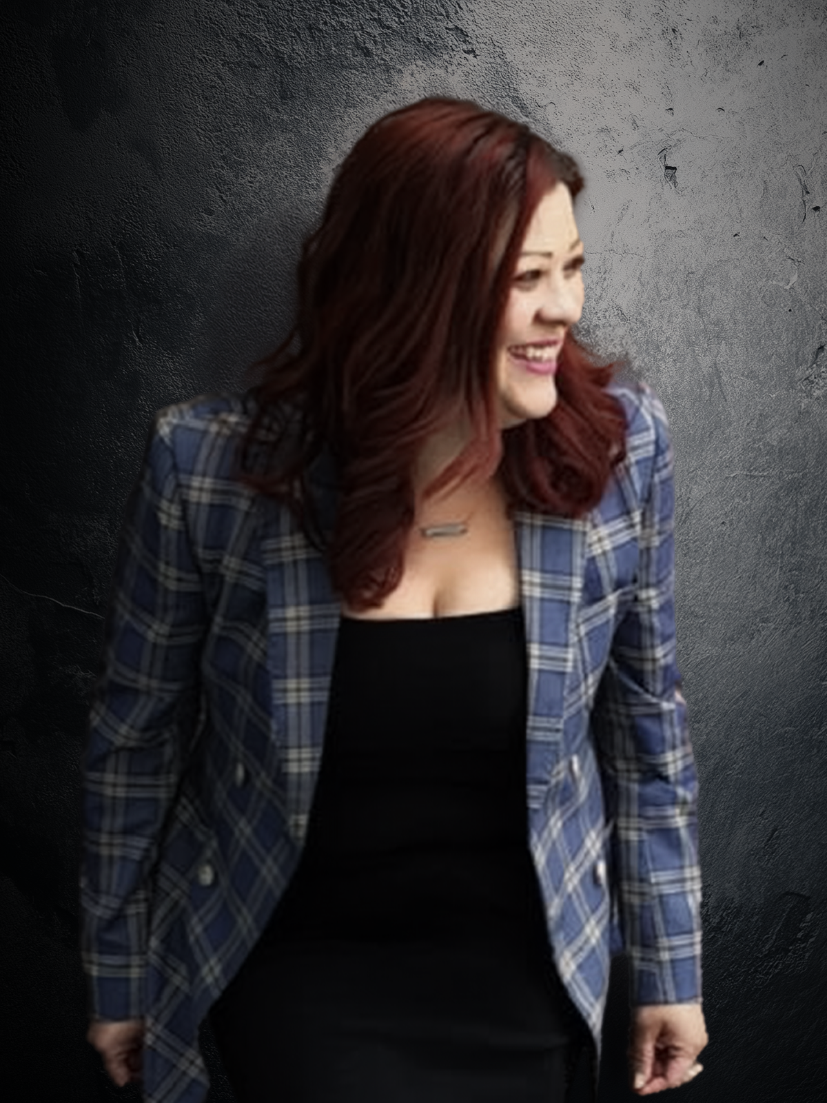 A woman with red hair smiling and looking to her right, wearing a blue plaid blazer over a black top, against a textured gray background.